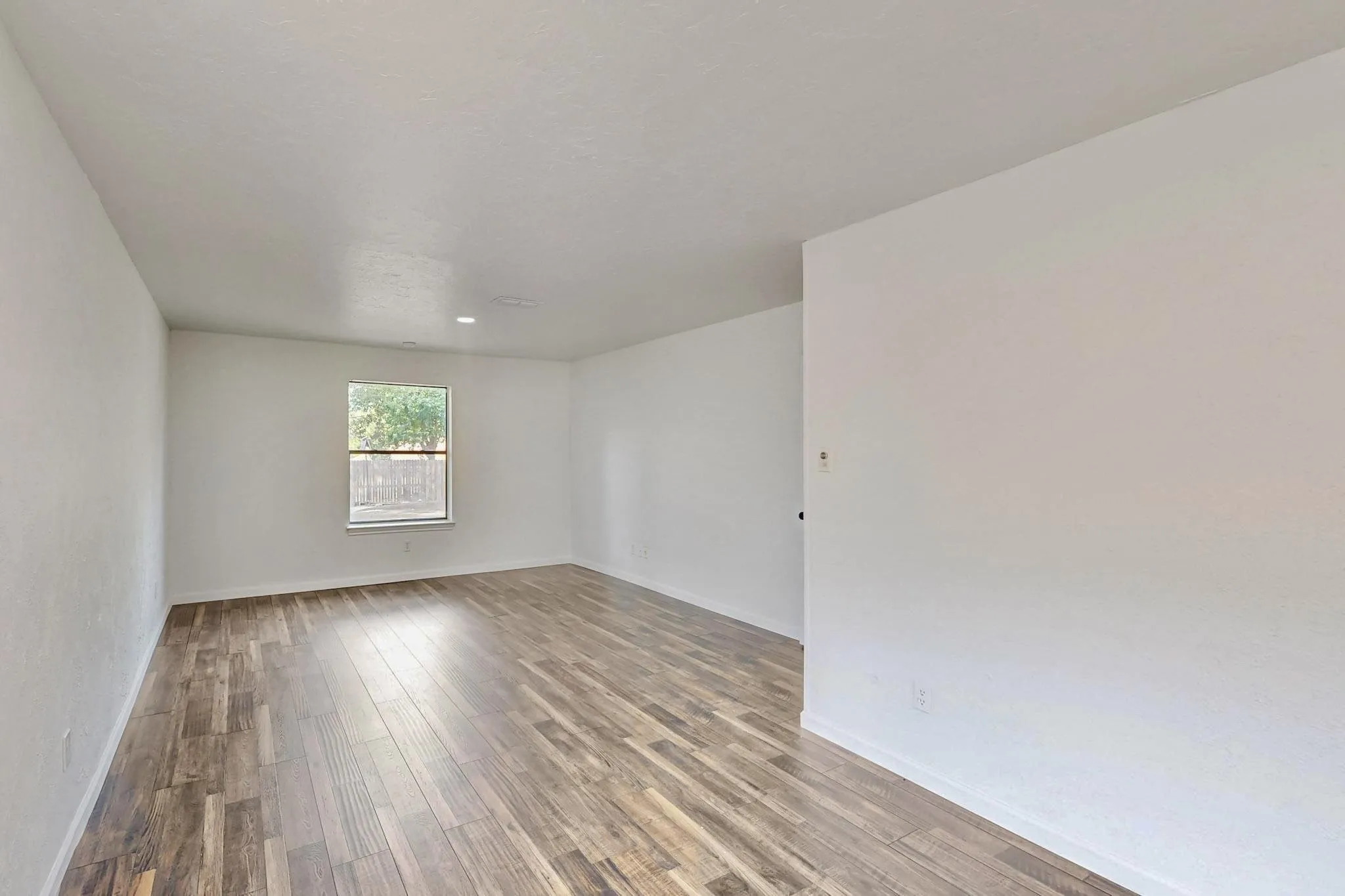 Bright interior space featuring wood-finish flooring, a single window with a framed outdoor scene, white painted walls, and recessed ceiling lighting