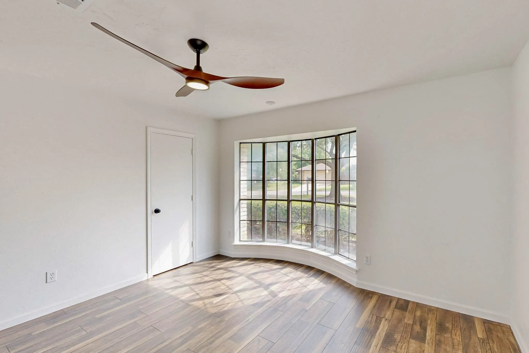 Room featuring a curved bay window with grid-patterned panes, wood-finish flooring, a modern ceiling fan with integrated lighting, a white door with a black handle, and white baseboards
