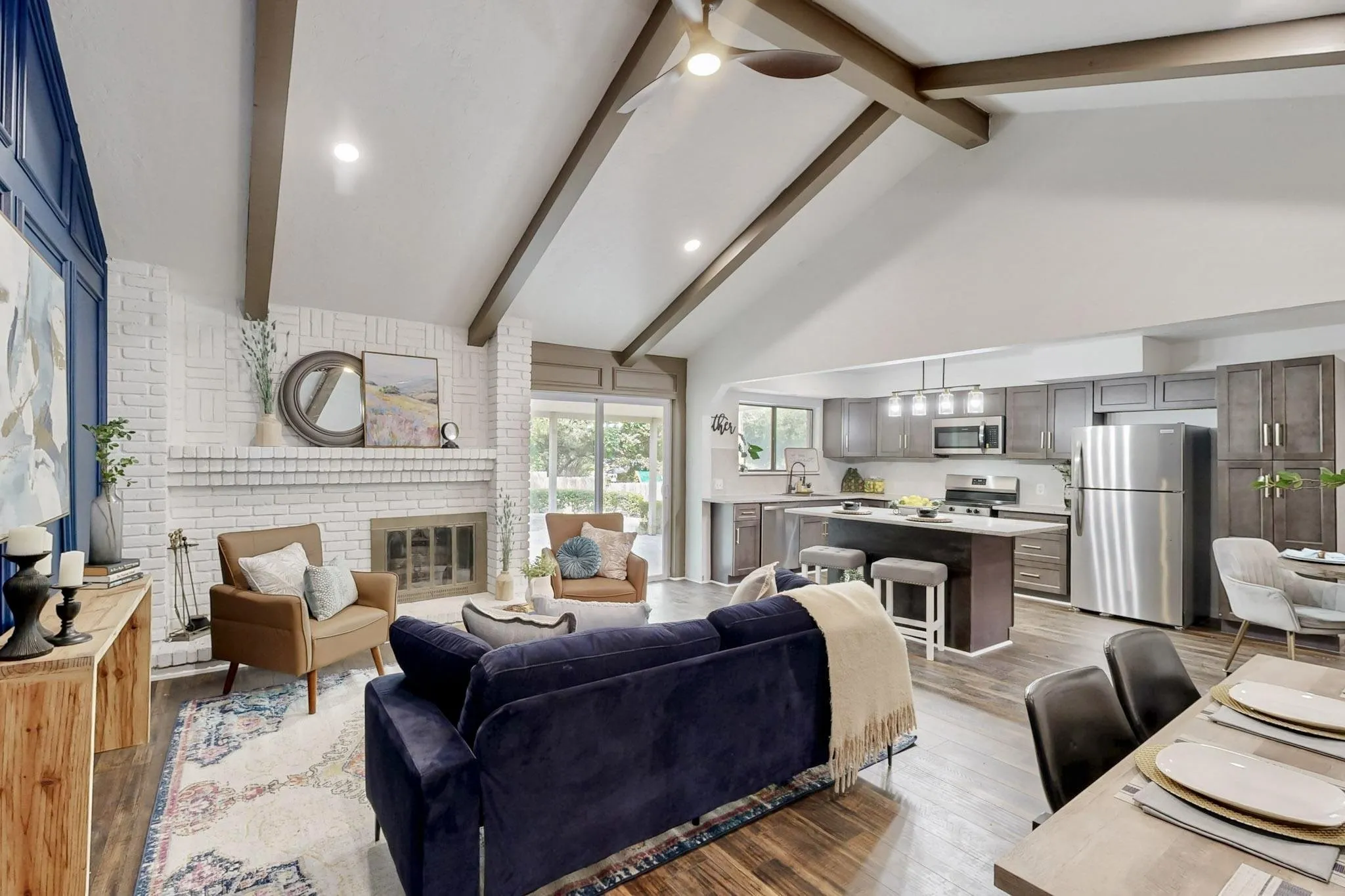 Vaulted ceilings with exposed beams, a white-painted brick fireplace, and wood-finish flooring define this open-concept living space