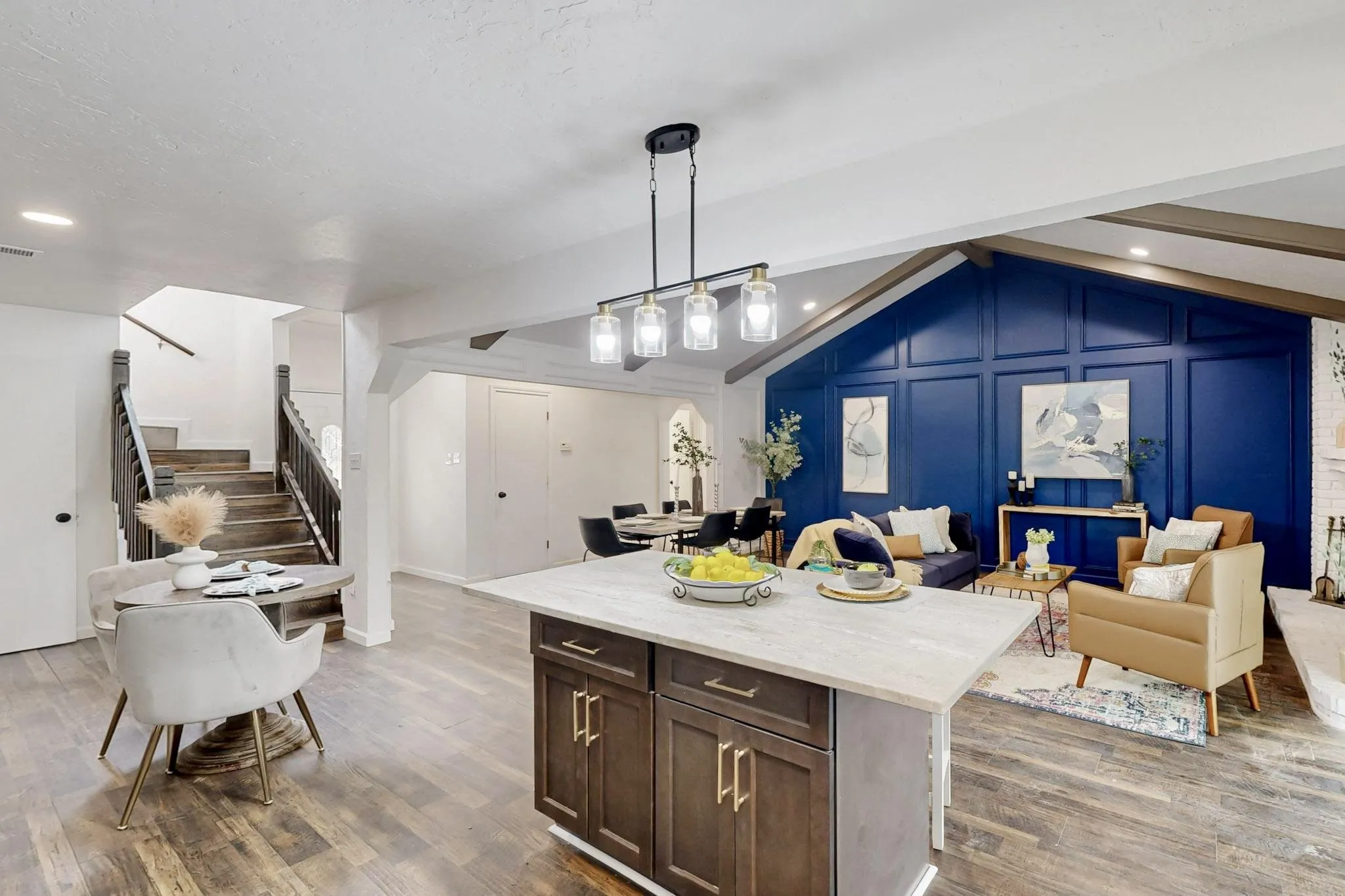 Open-concept living space with wood-finish flooring, a kitchen island featuring a light-toned countertop, a vaulted ceiling with exposed beams, and a striking blue accent wall with decorative paneling