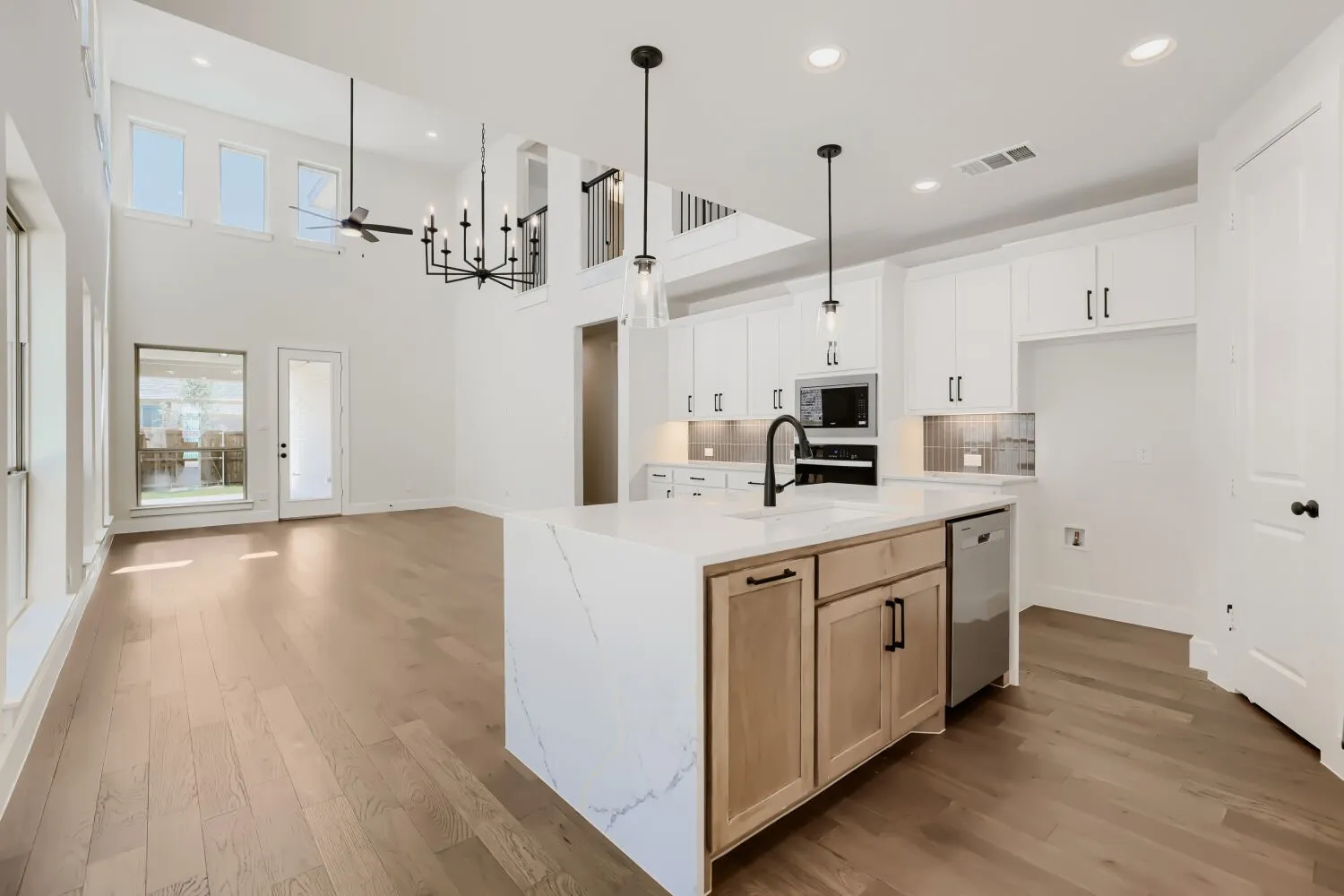Open-concept main living space featuring a central kitchen island with waterfall countertop, wood-finish flooring, two-story ceilings with multiple windows, and a modern black chandelier