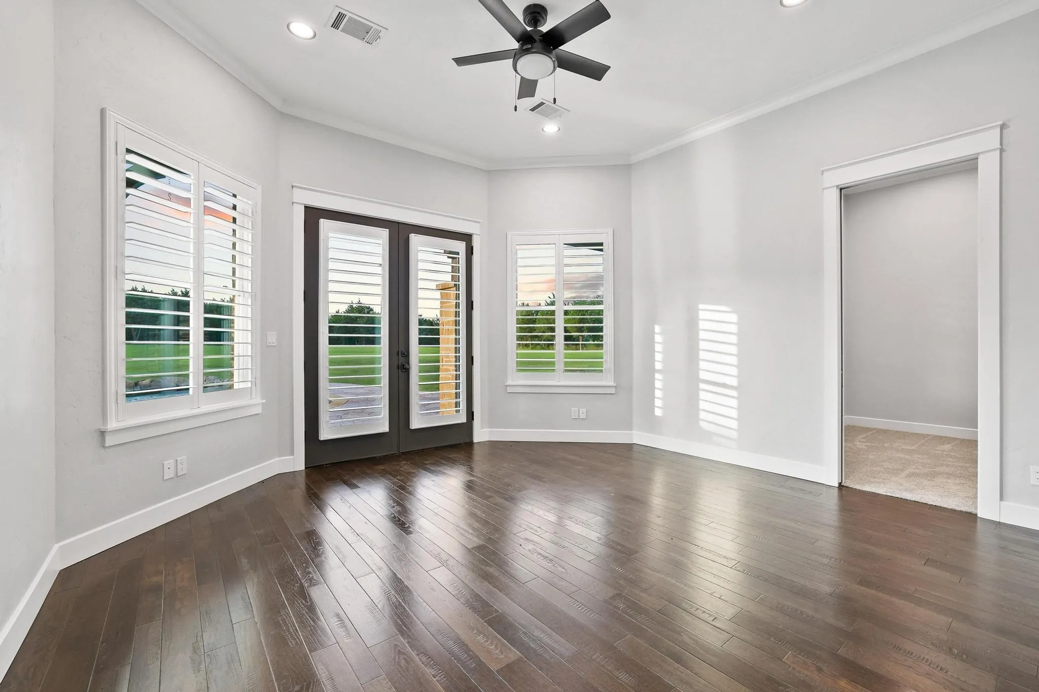 Spacious room featuring rich wood-finish flooring, white plantation shutters, and a modern ceiling fan
