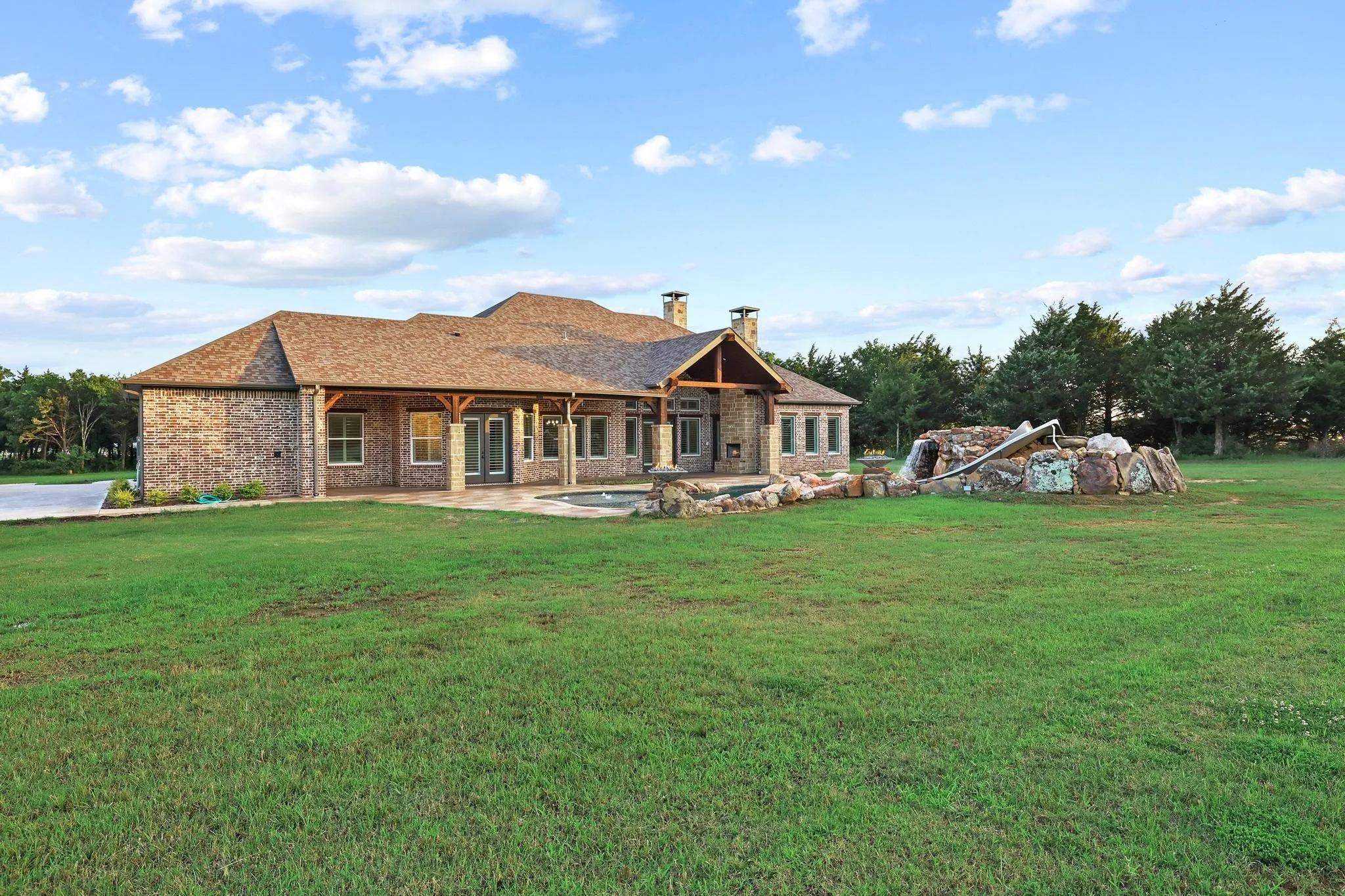 Spacious backyard featuring a brick exterior home, covered patio with wood beam accents, a swimming pool, and a stone-built grotto with a slide
