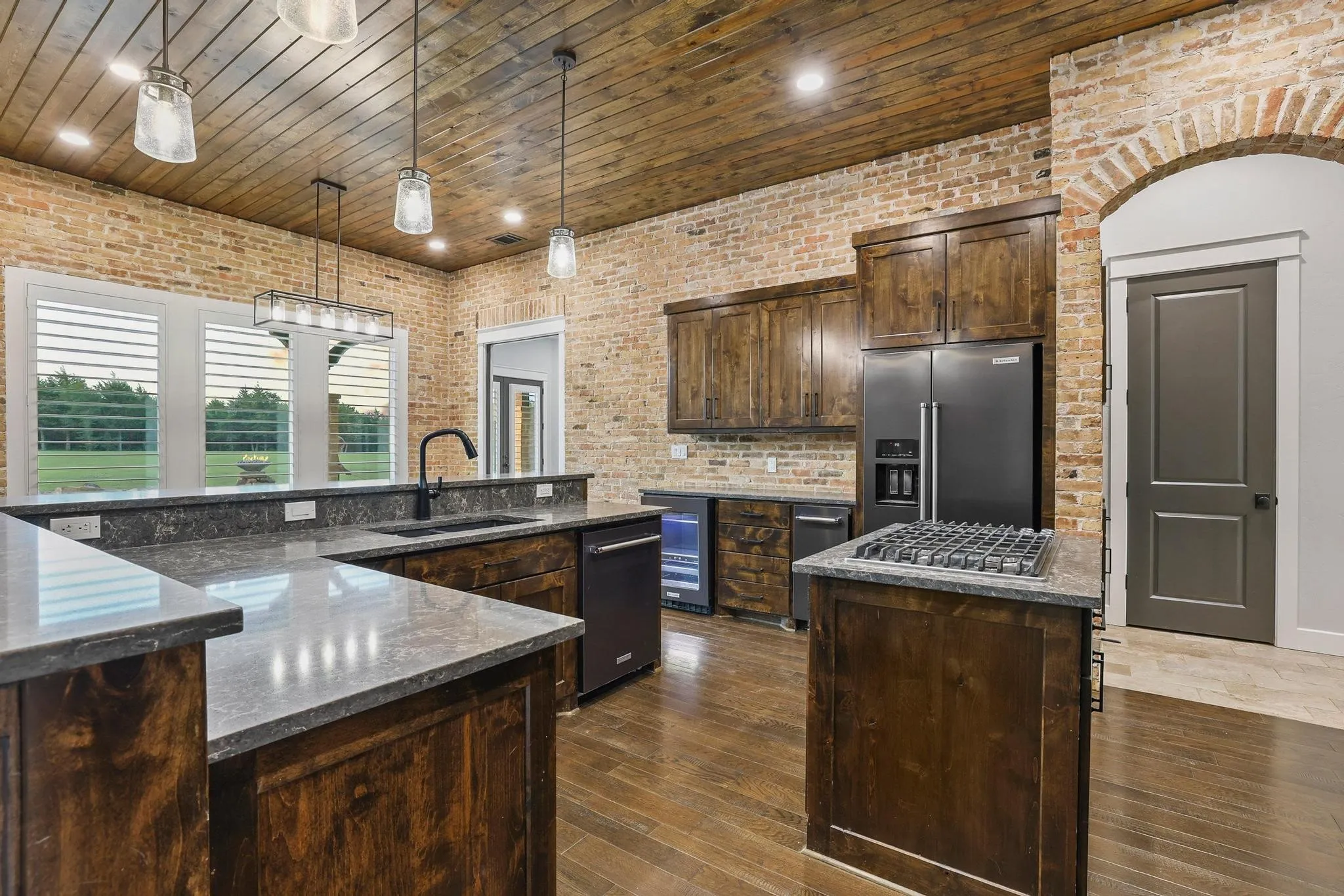 Kitchen featuring exposed brick walls, wood-finish flooring, dark wood cabinetry, and stone countertops