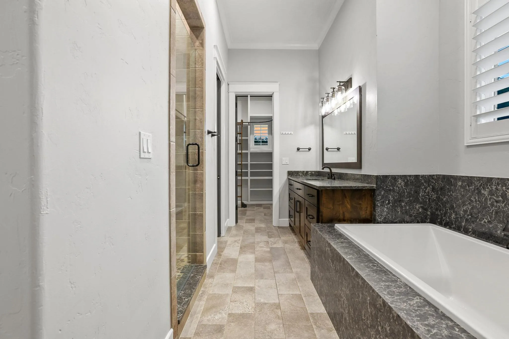 Bathroom featuring a built-in soaking tub with stone surround, a wood-finish vanity with stone countertop and backsplash, glass-enclosed shower, tile flooring, and integrated shelving