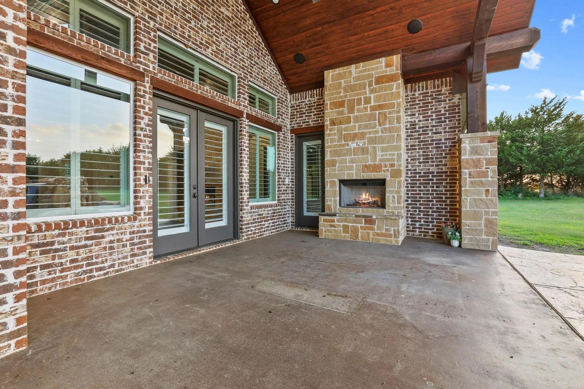 Covered outdoor patio featuring a stone fireplace, exposed wood-beam ceiling, brick exterior, and French doors with privacy shutters