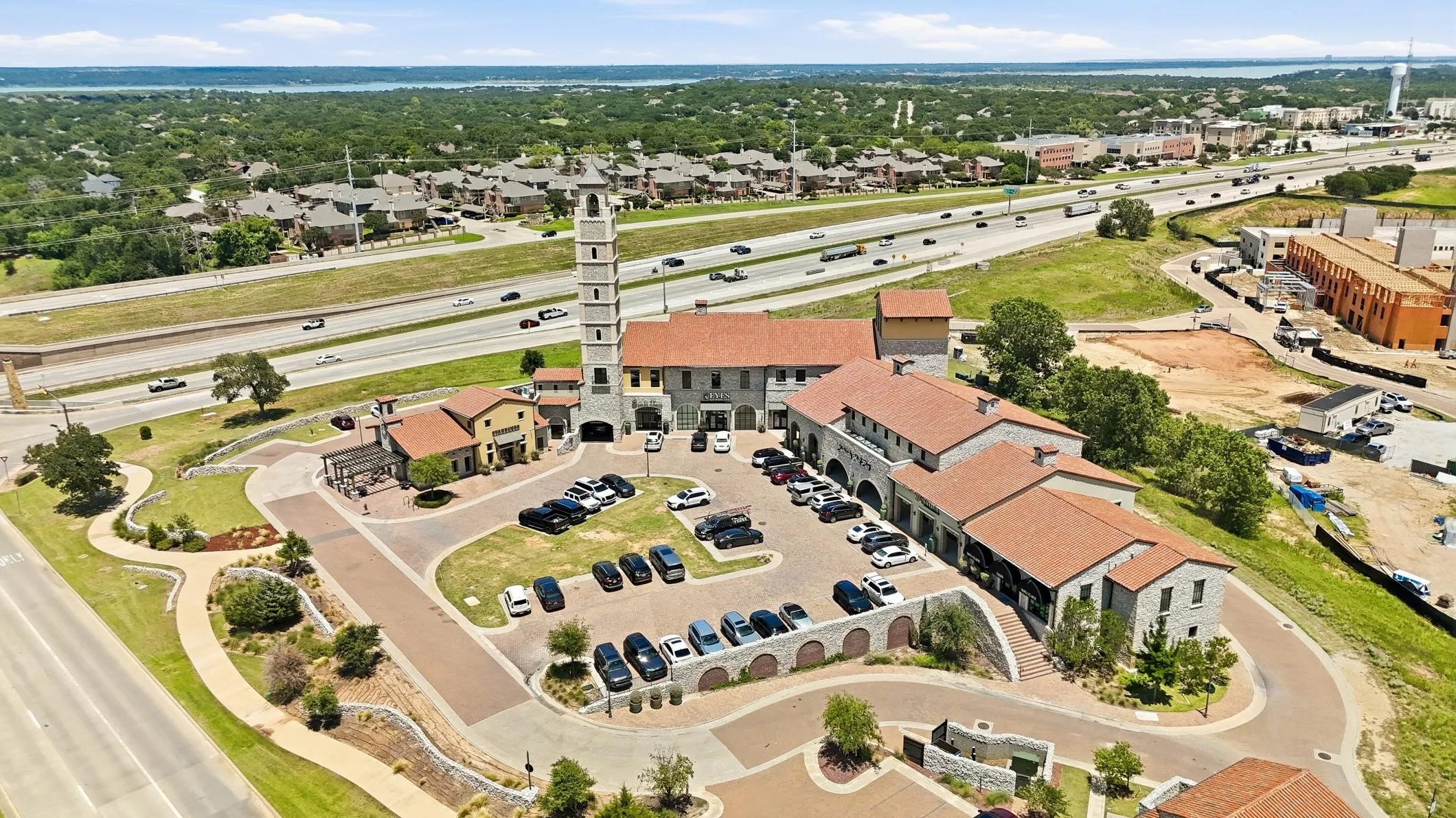 Mixed-use development featuring a prominent bell tower, terracotta-style roofing, stone-clad facades, multiple storefronts, and a visible surface parking area