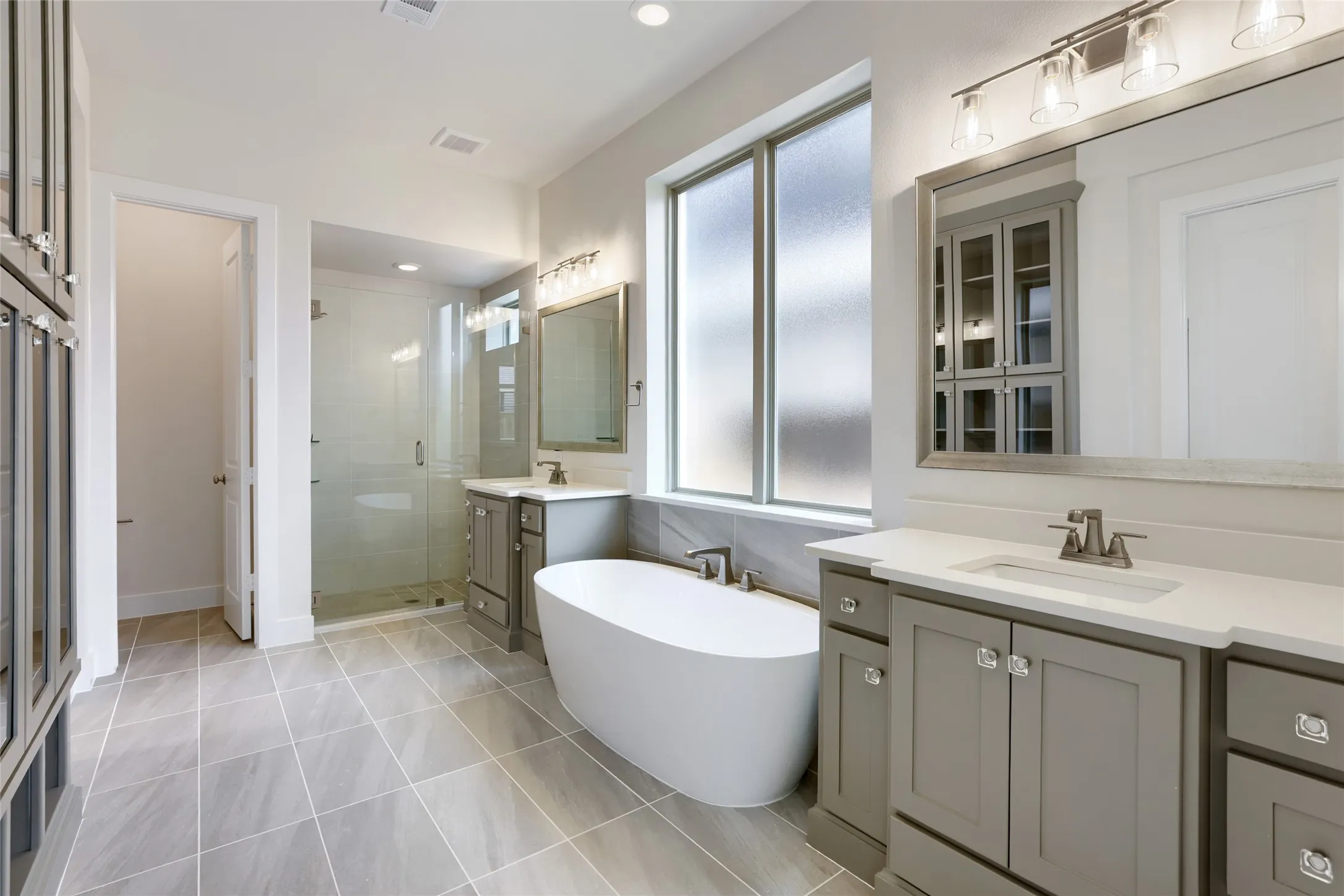 Spacious bathroom featuring a freestanding soaking tub, dual vanity with white countertops, expansive glass-enclosed shower, large frosted windows, and large format tile flooring