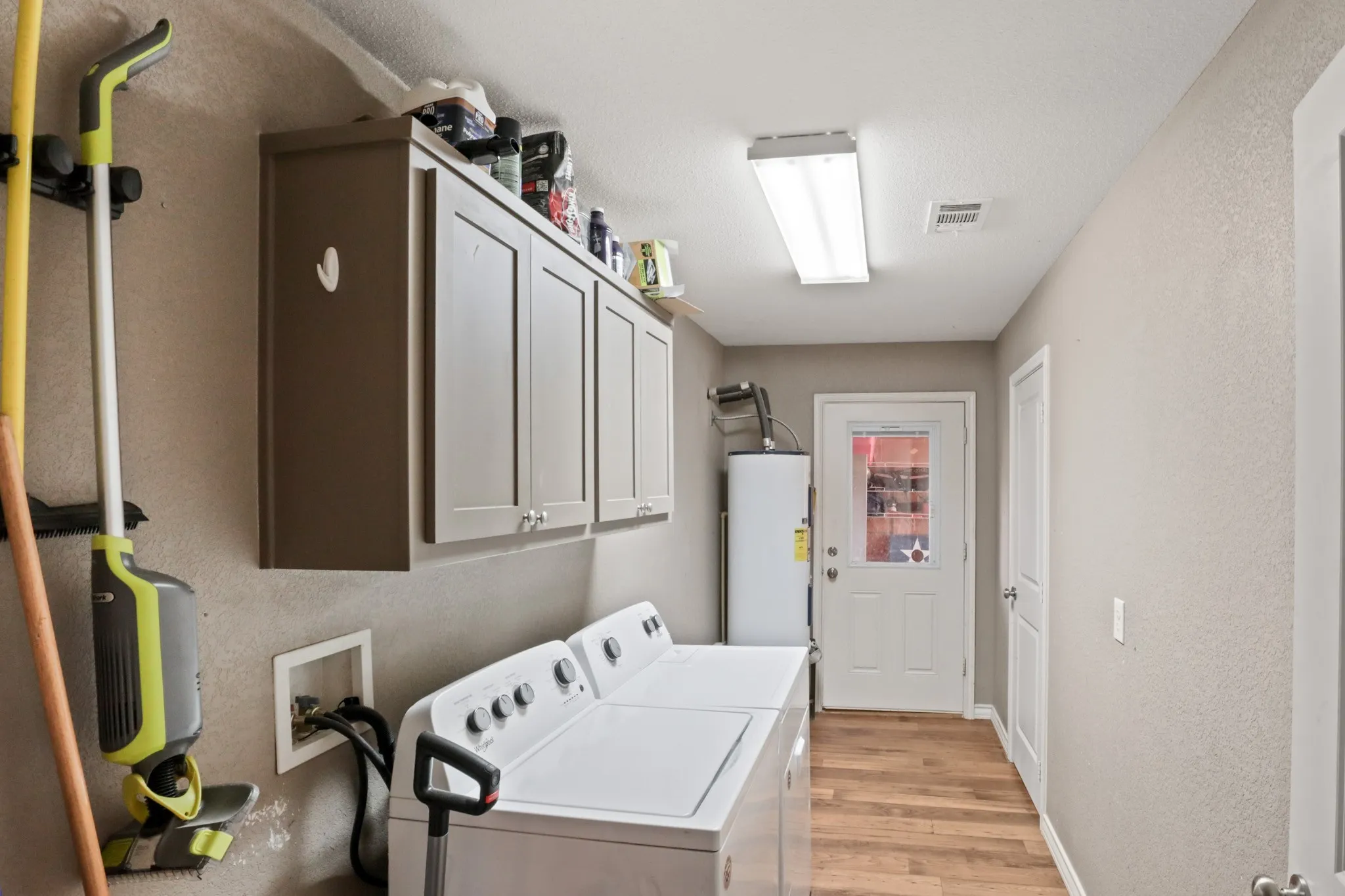 Utility room featuring wood-finish flooring, overhead fluorescent lighting, and built-in upper cabinetry with silver-toned hardware
