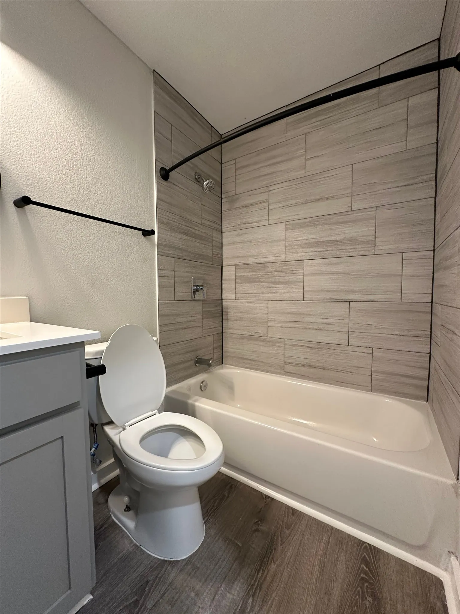 Bathroom featuring a built-in tub with a tile surround, coordinating wood-finish flooring, a white vanity countertop, and dark-finish fixtures