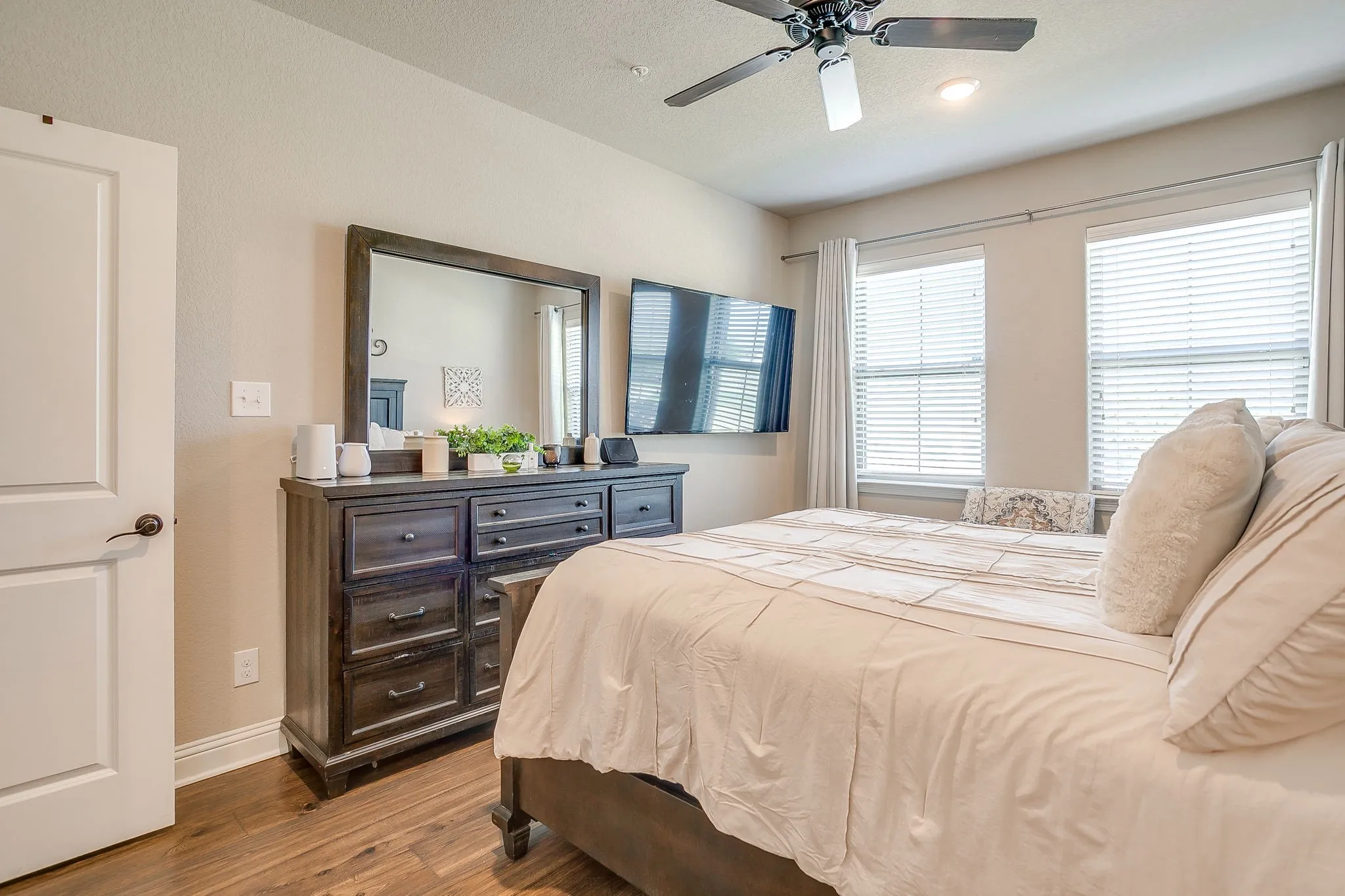 Spacious room featuring wood-finish flooring, two large windows with blinds, a ceiling fan with integrated lighting, a built-in wall-mounted television, and a white interior door with dark bronze hardware