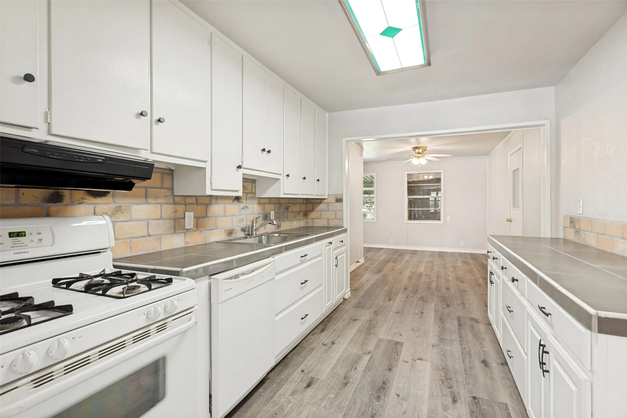Galley kitchen featuring white cabinetry, brick-finish backsplash, dark countertops, wood-finish flooring, and integrated white appliances