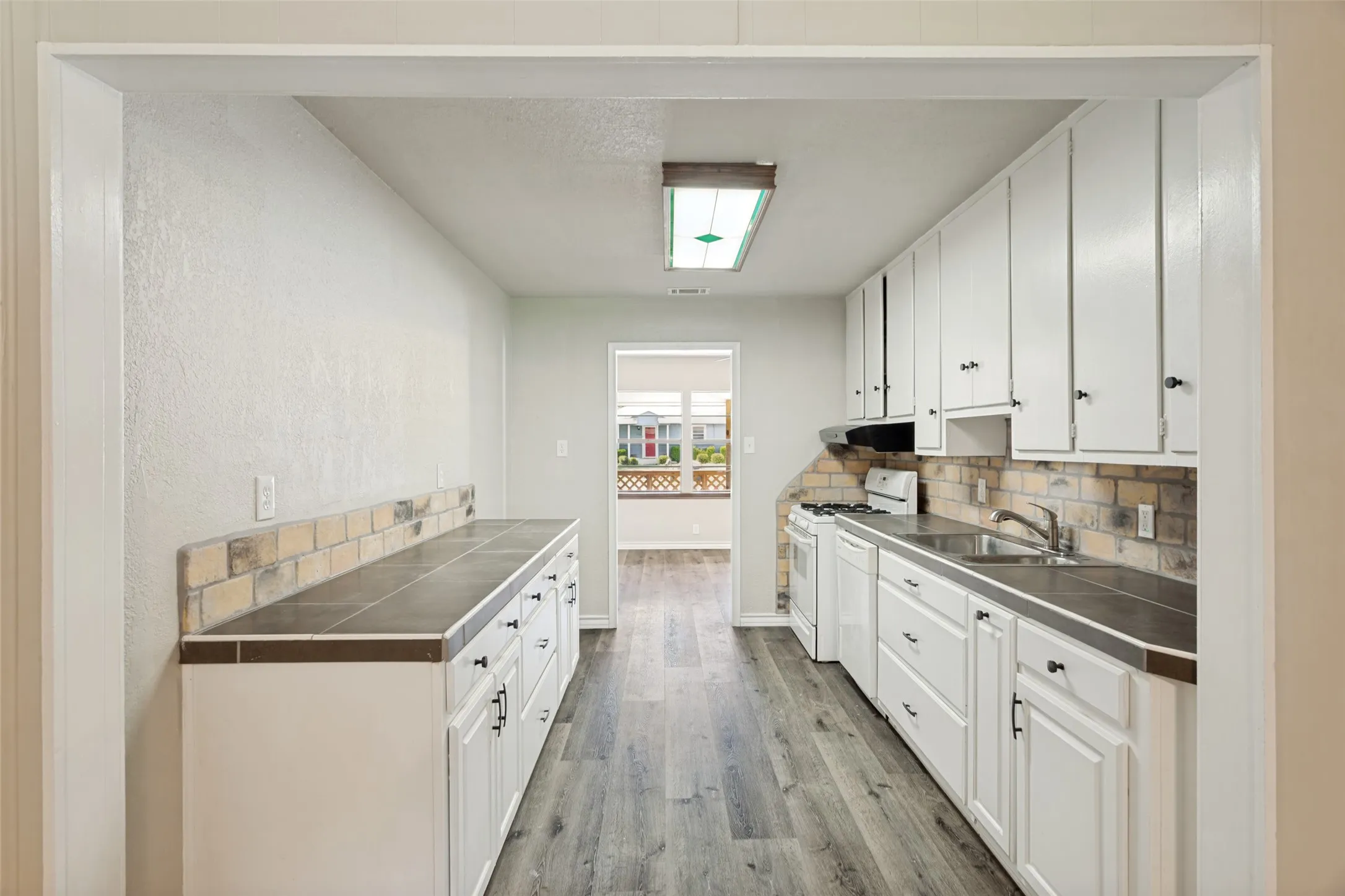 Galley kitchen featuring white cabinetry, tile countertops, a brick-style backsplash, wood-finish flooring, and a flush-mount ceiling light fixture