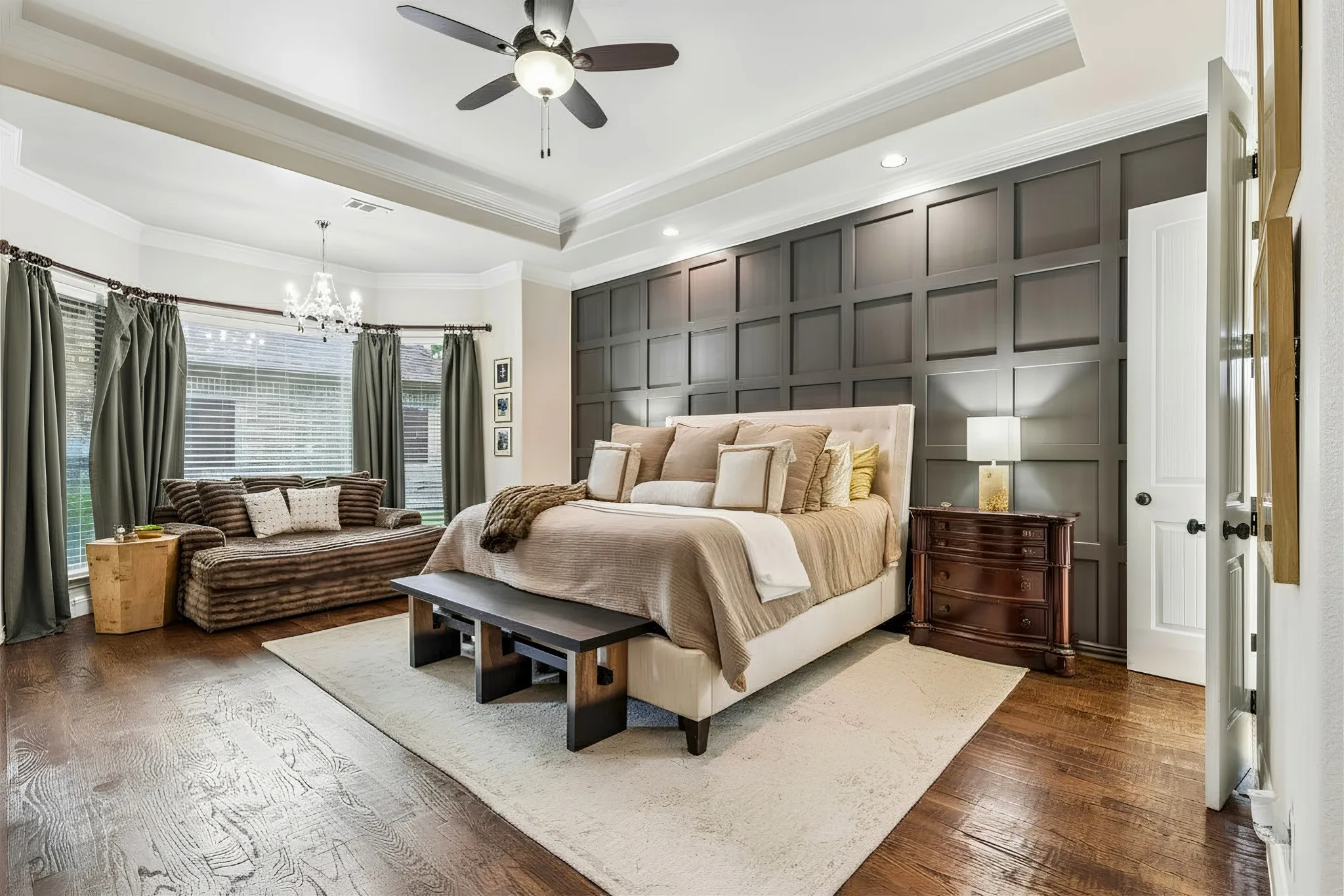 Dark wood-finish flooring, accent wall with wainscoting, tray ceiling, ceiling fan, and bay window with blinds