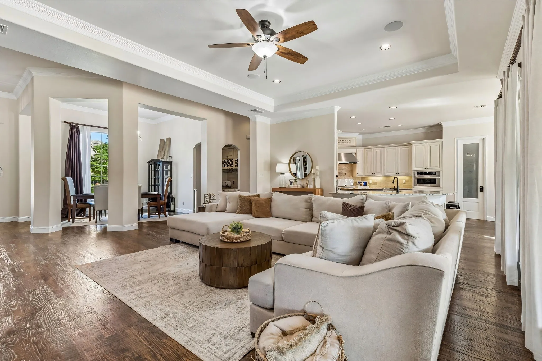Wood-finish flooring throughout, tray ceiling with a built-in ceiling fan, recessed lighting, and an open layout connecting to the kitchen featuring light-toned cabinetry and stainless steel appliances