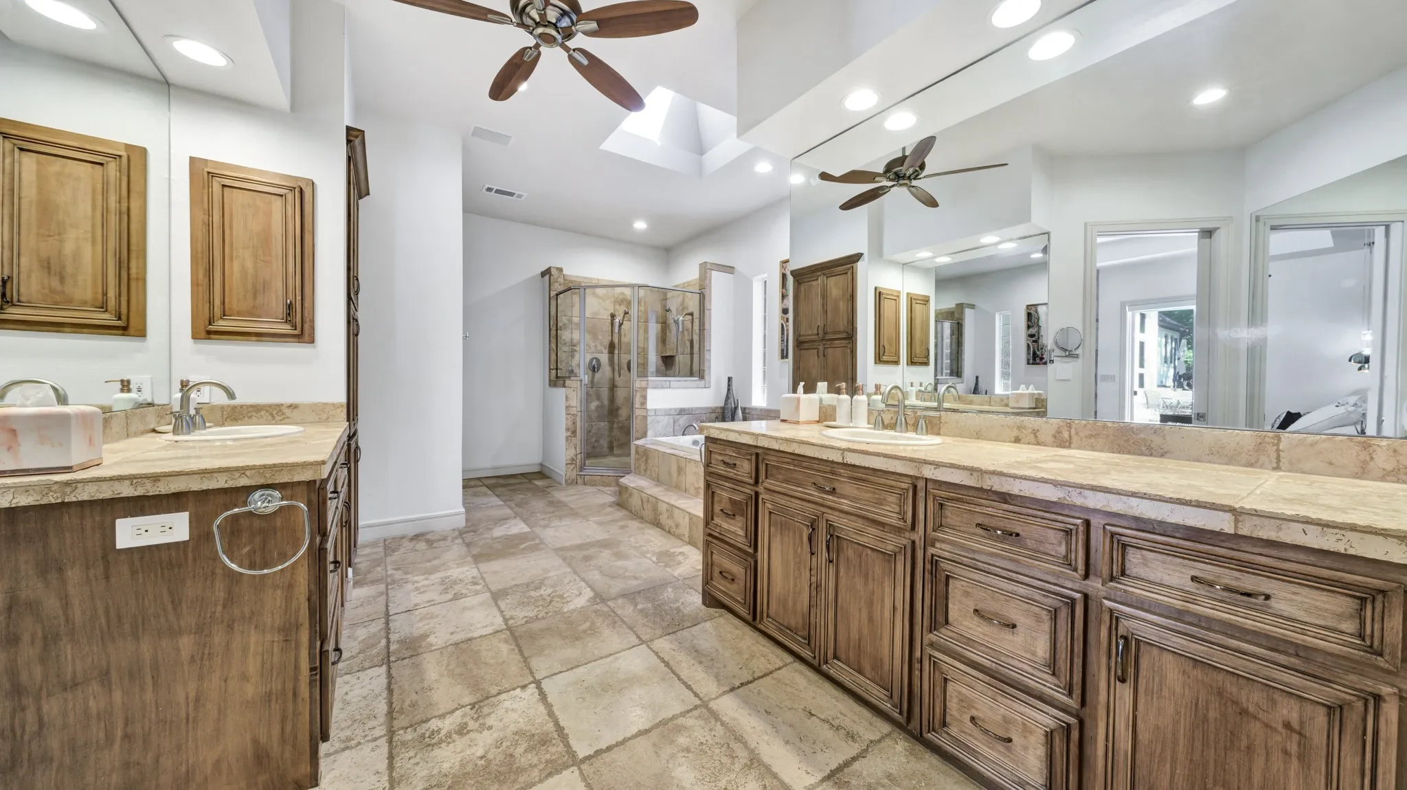 Spacious bathroom featuring twin wood-finish vanities with light stone countertops, a skylight, a built-in jetted tub, and a separate stone-tiled shower enclosure
