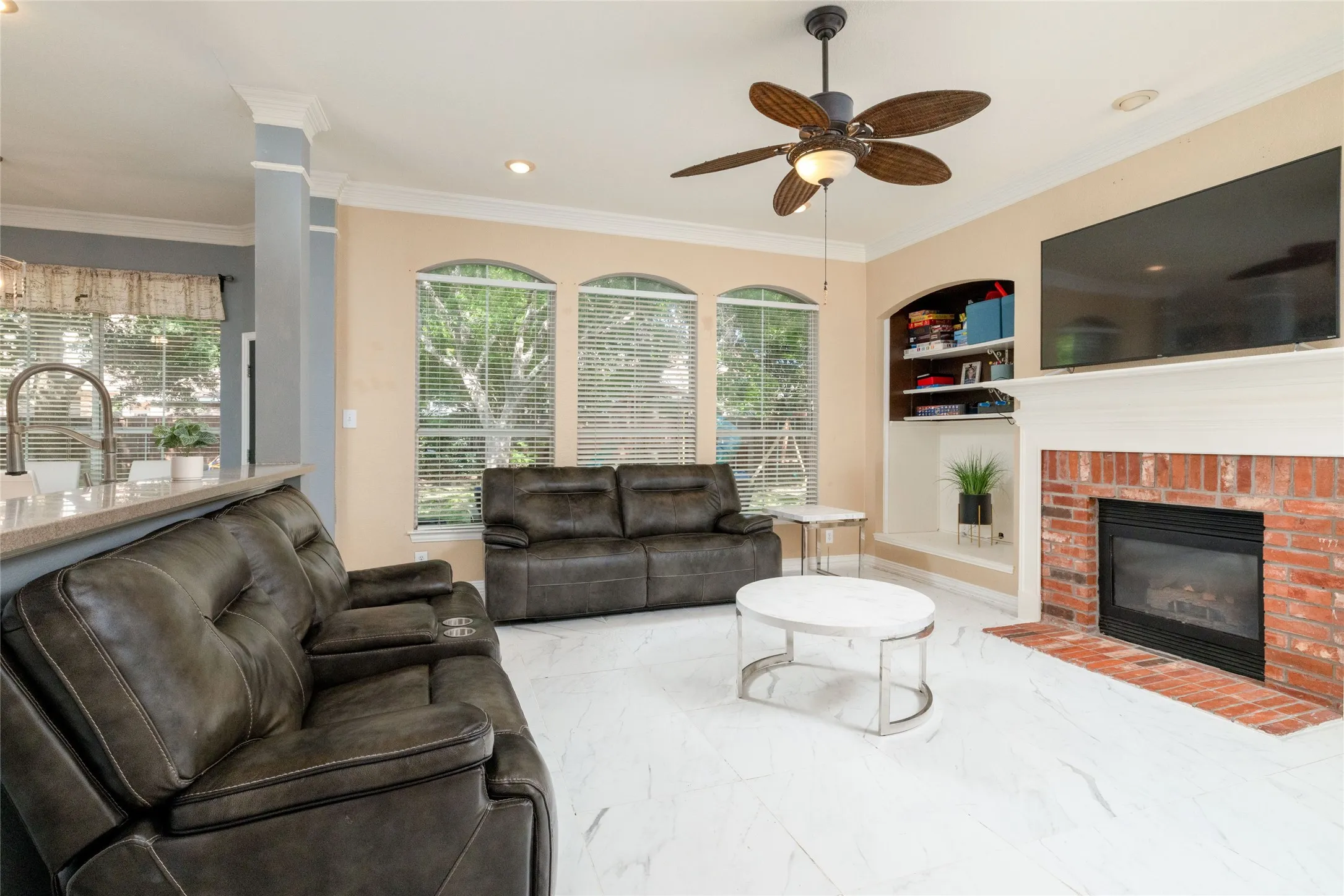 Living area featuring large format white tile flooring, a brick fireplace with white mantel, a built-in shelving unit, and a ceiling fan with integrated lighting