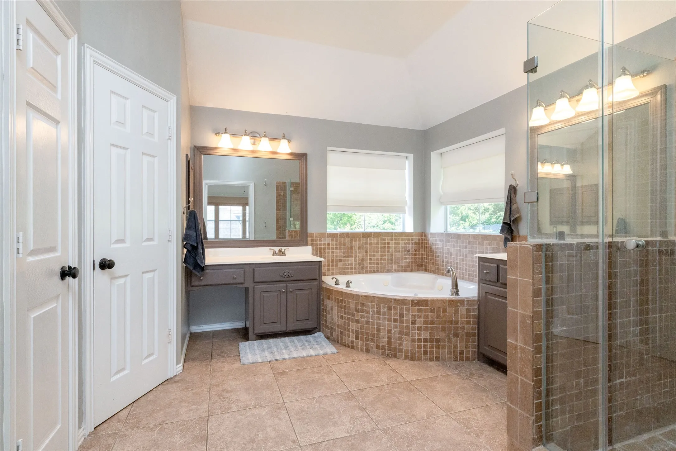 Spacious bathroom featuring an oval jetted tub with tile surround, a glass-enclosed shower, dual vanities with integrated sinks and stone countertops, and tile flooring