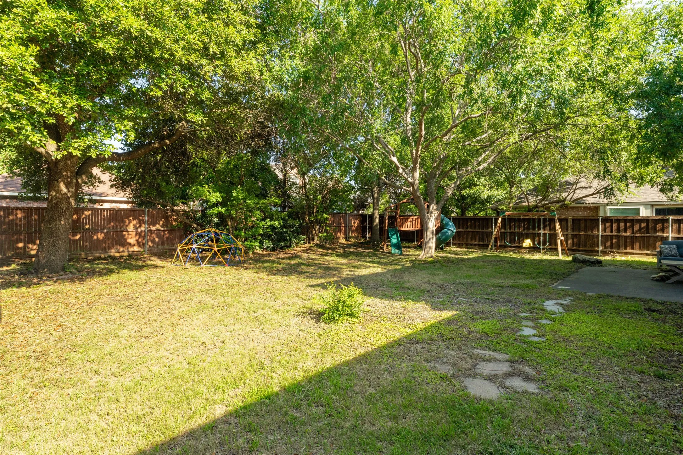 Expansive lawn area featuring mature trees providing natural shade, a wood privacy fence, and stepping stone path