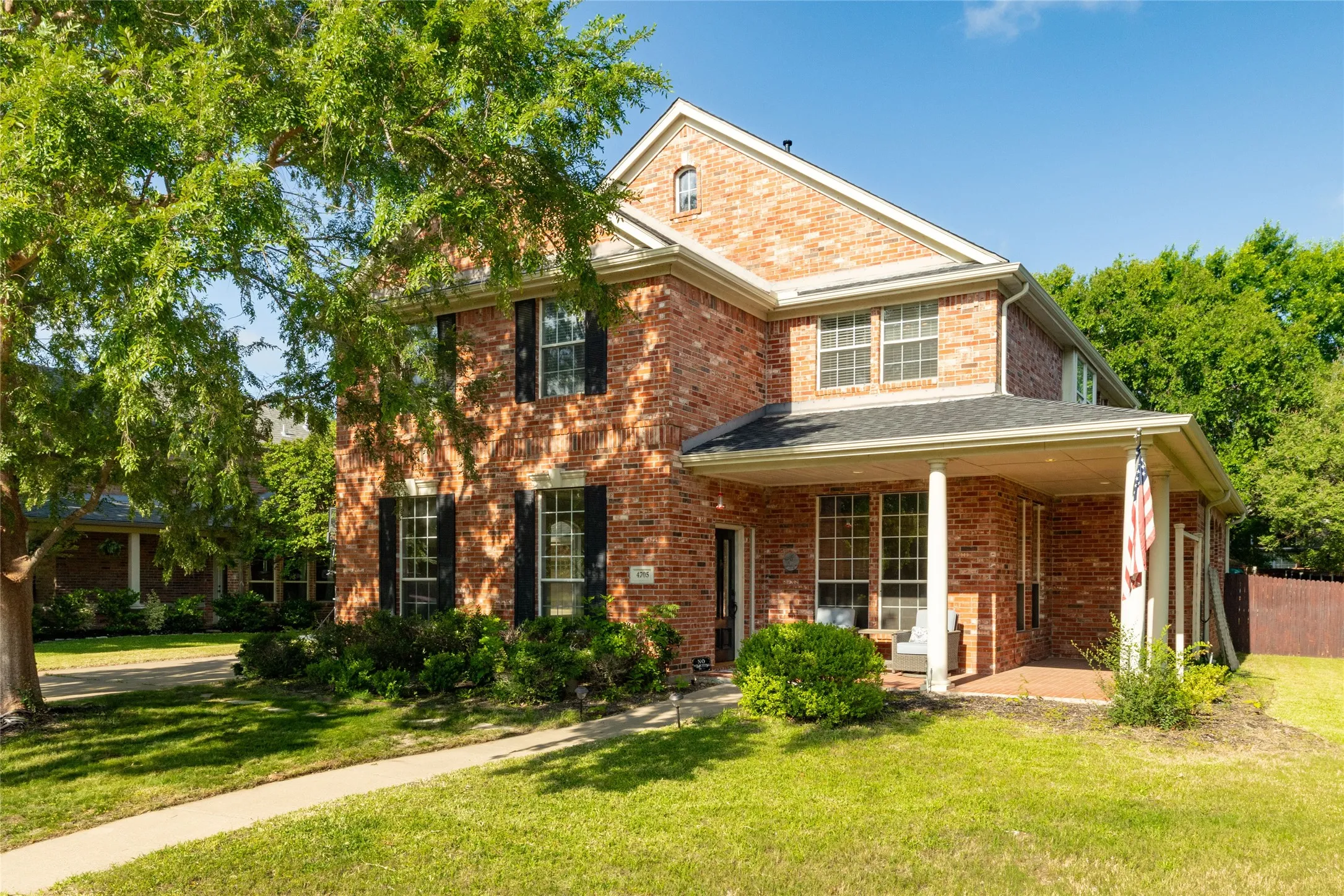 Brick facade residence featuring a covered front porch with white columns, black louvered shutters, and mature trees