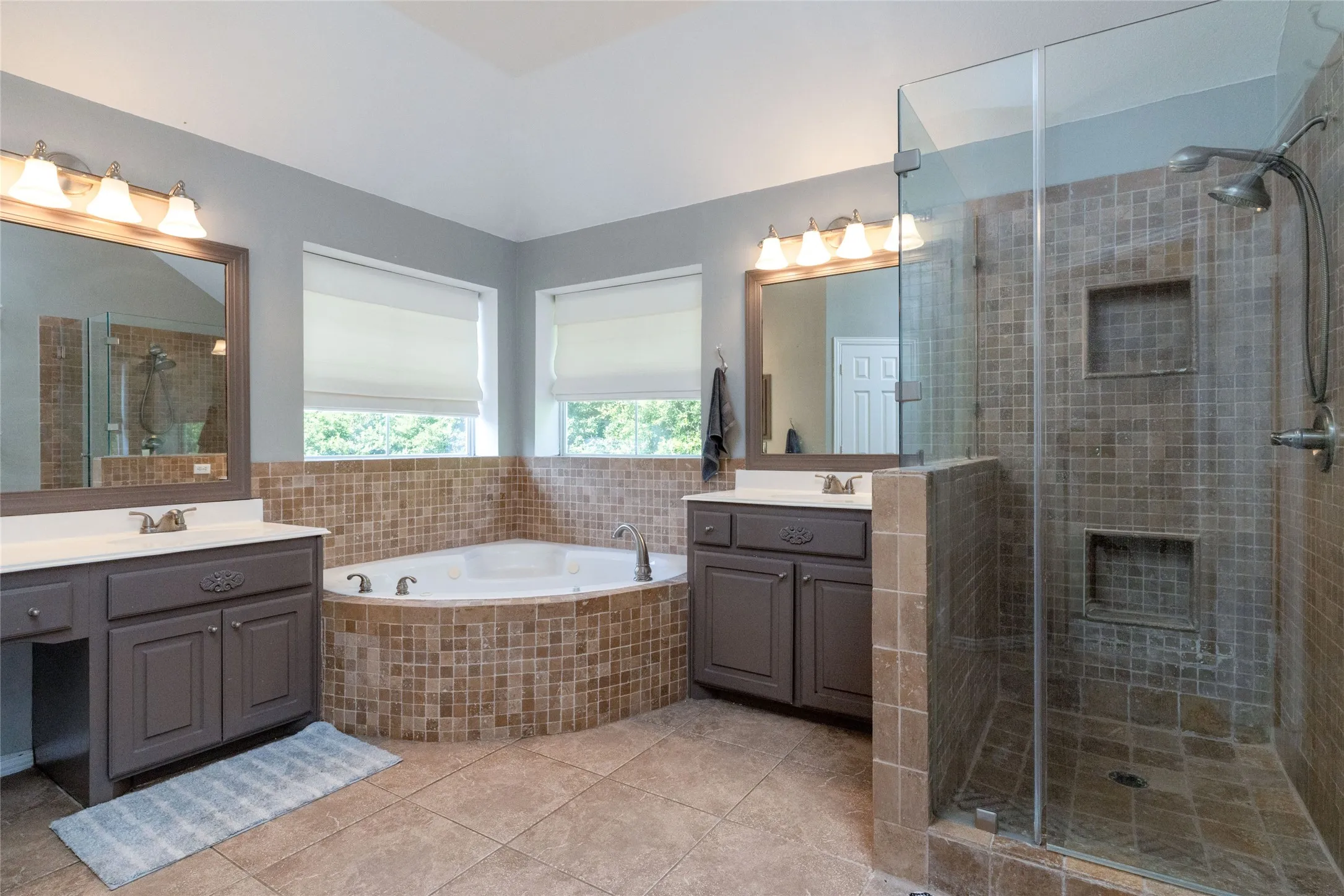 Bathroom featuring a corner jetted tub with mosaic tile surround, a glass-enclosed shower with dual showerheads and recessed shelving, and two individual vanities with integrated sinks and storage cabinets