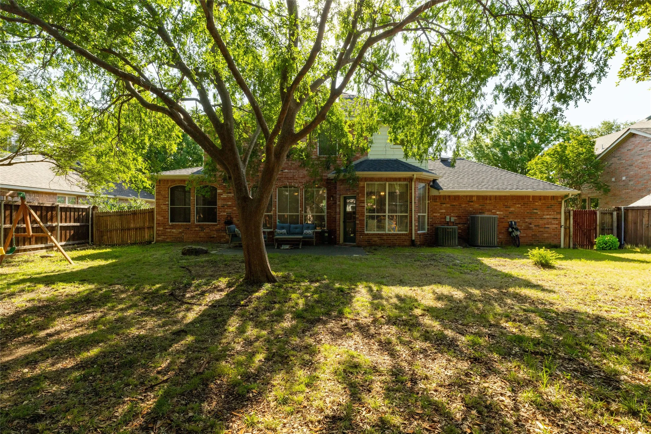Spacious backyard featuring a substantial mature tree, brick exterior, and a partially covered patio area
