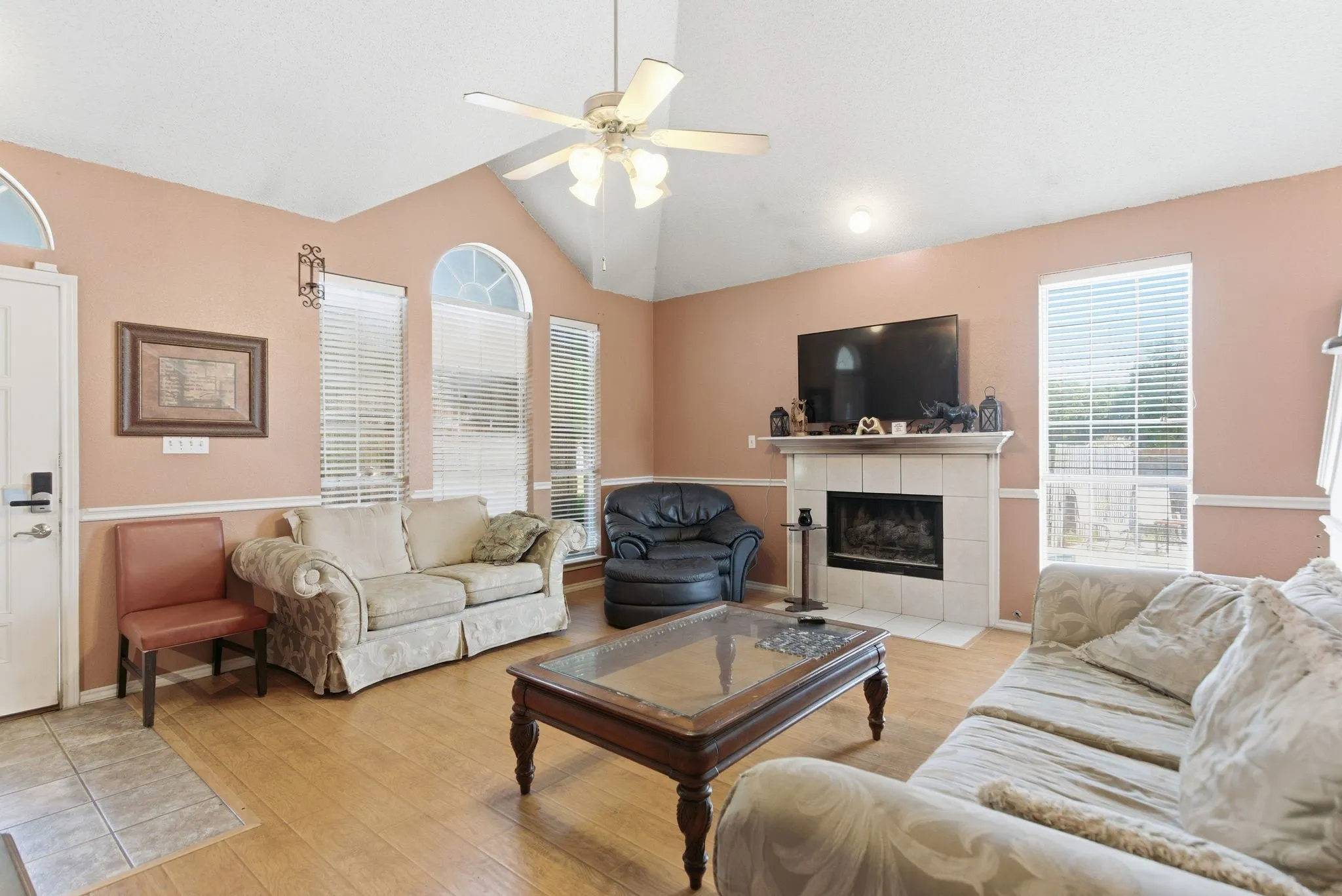 Spacious living area featuring vaulted ceilings, wood-finish flooring, a tiled fireplace with a mantel, arched windows, and a ceiling fan with integrated lighting