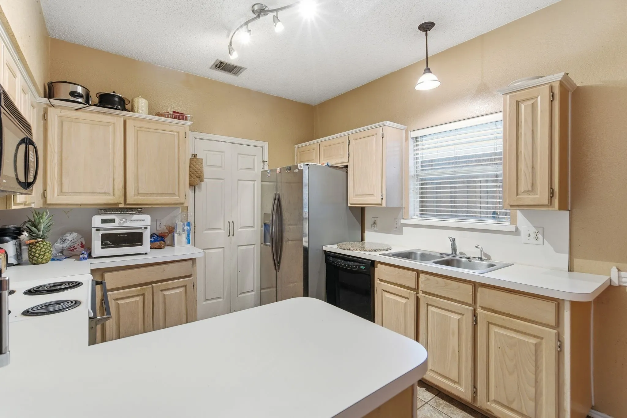 Kitchen featuring a central island with a white countertop, light wood-finish cabinetry, a double basin sink, and a window with blinds