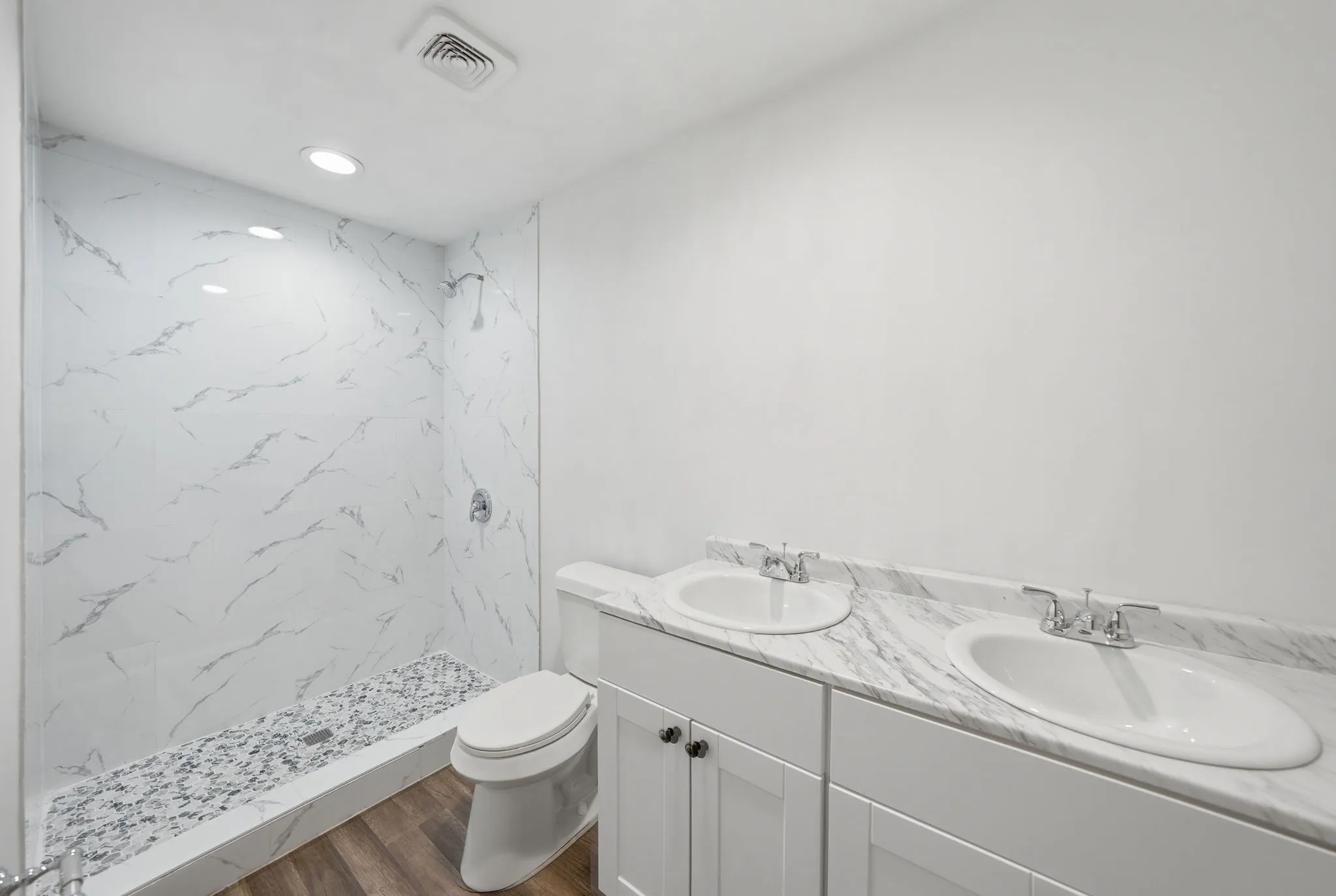 Bathroom featuring a walk-in shower with white marbled tile walls and a pebble floor, a dual vanity with white marbled countertops and integrated sinks, white shaker-style cabinetry with dark hardware, and wood-finish flooring