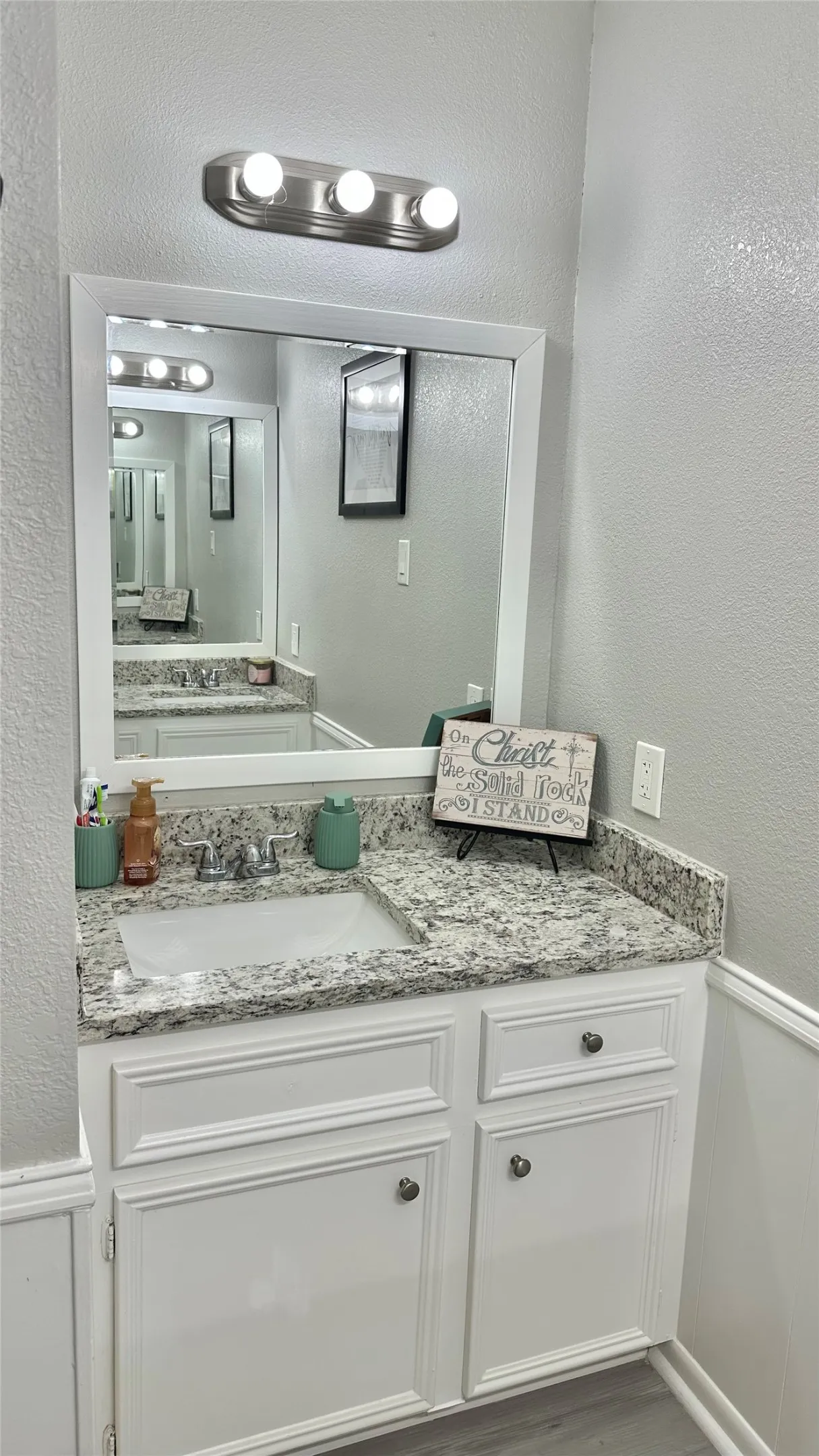 Bathroom vanity with a white framed mirror, brushed nickel light fixture, granite countertop, undermount sink, and white cabinetry featuring decorative paneling