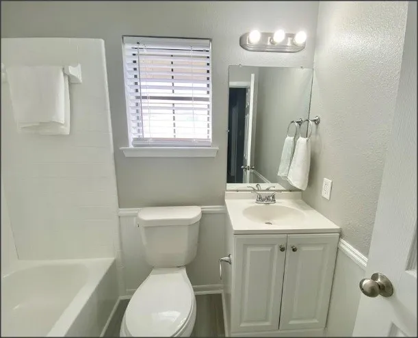 Full guest hallway bathroom featuring a white porcelain tub with integrated shower, single-pane window with blinds, white toilet, and a white vanity with integrated sink