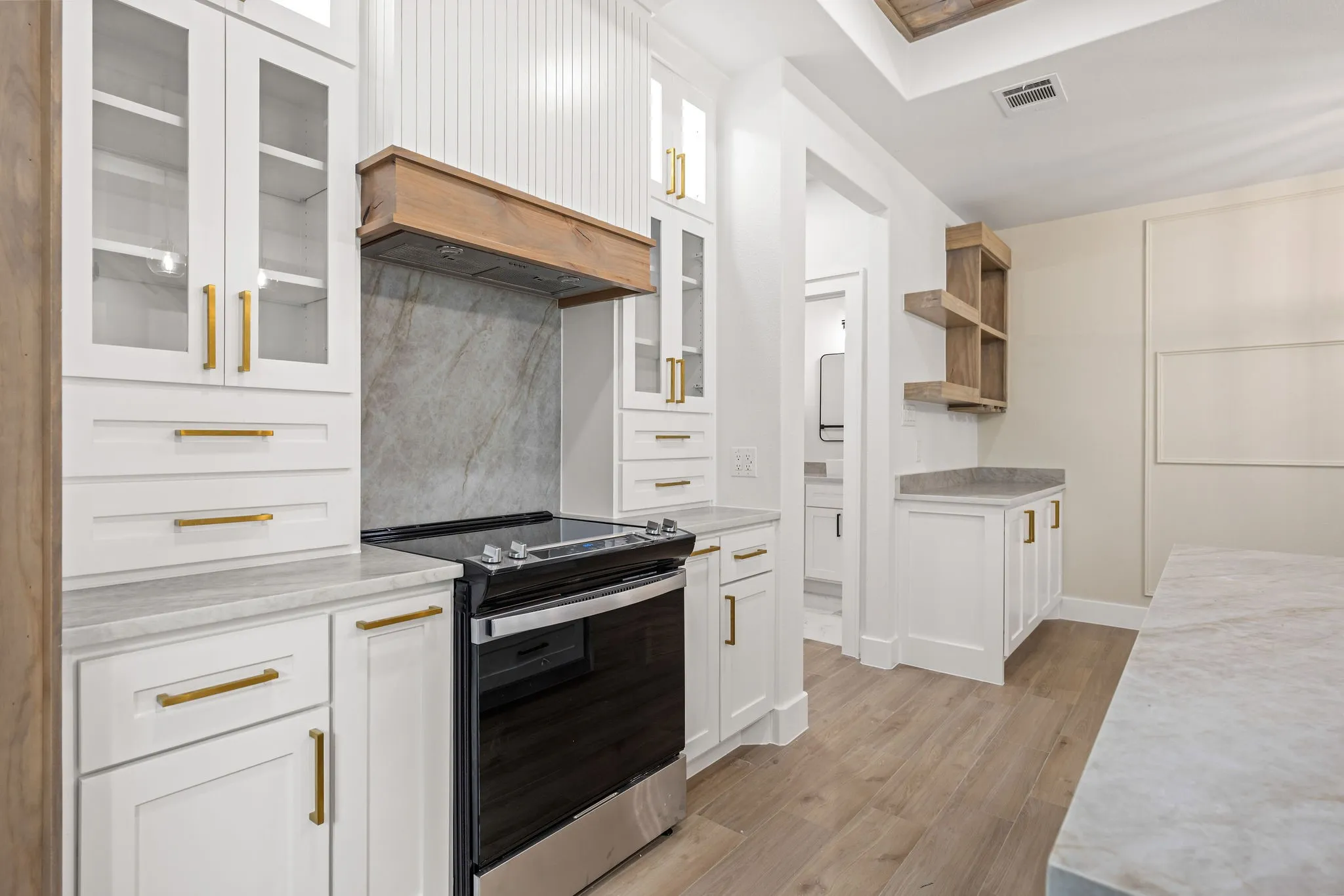 Contemporary kitchen featuring white cabinetry with gold-finish hardware, a built-in stainless steel range, and a wood-finish range hood
