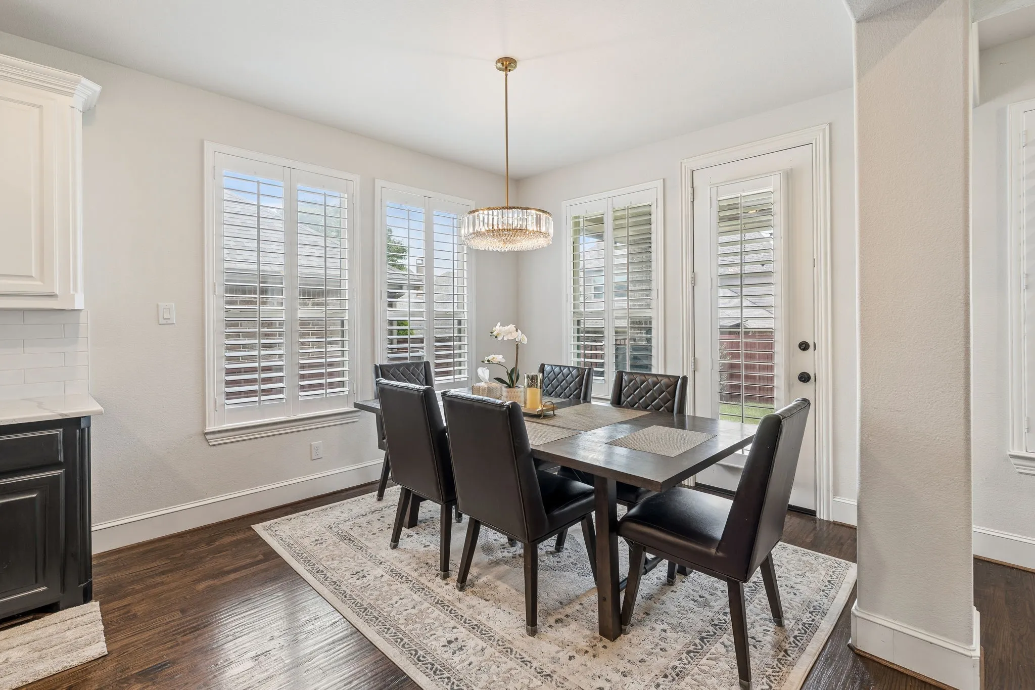 Dining area featuring wood-finish flooring, multiple windows with white plantation shutters, a glass-encased chandelier, and a white paneled door with black hardware