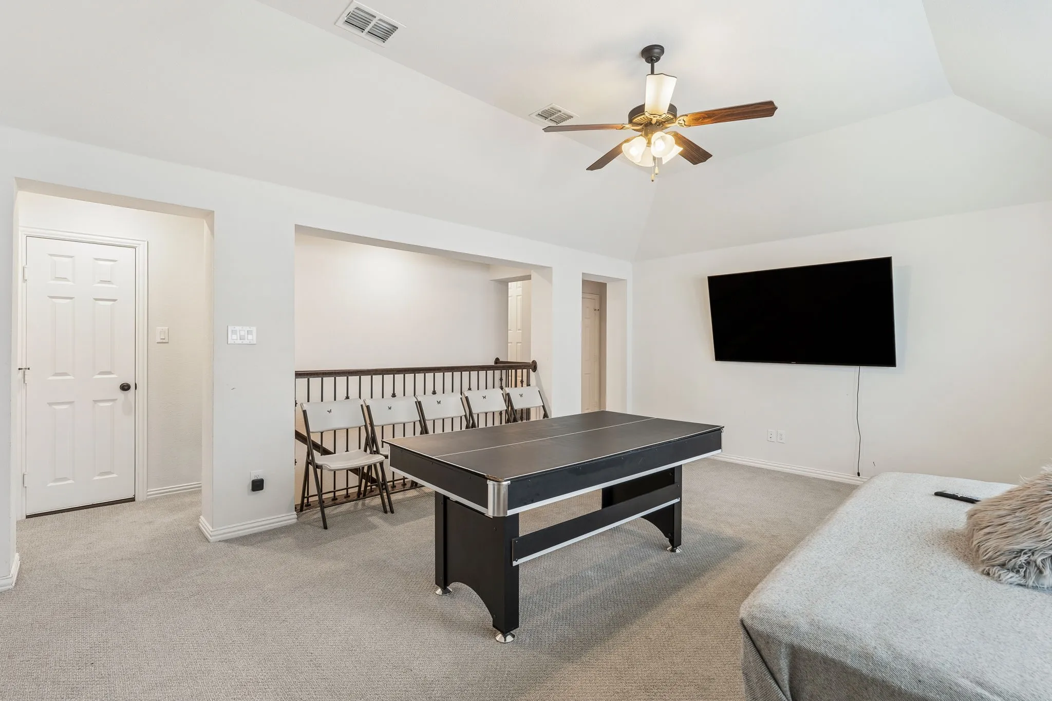 Carpeted entertainment space featuring a vaulted ceiling with a ceiling fan, wall-mounted television, and an interior balcony with wood balusters