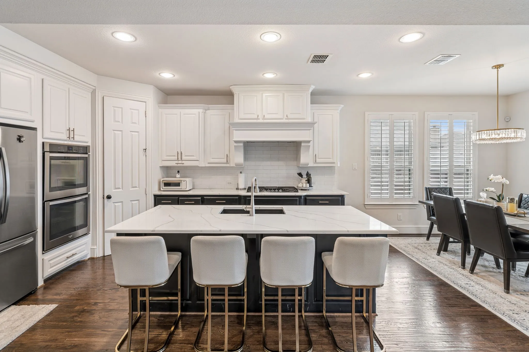 Kitchen featuring a spacious island with seating, dark wood-finish flooring, white cabinetry, integrated stainless steel double ovens, and a dining area with plantation shutters