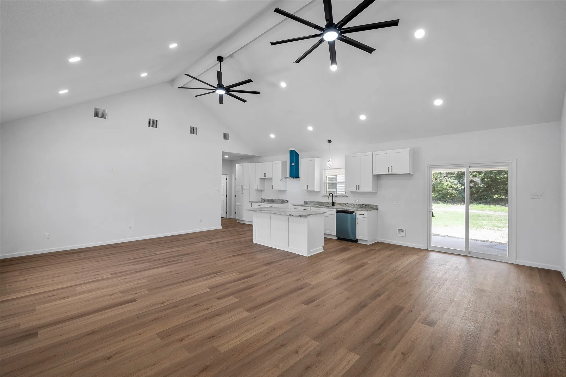Open-concept living space featuring vaulted ceilings with recessed lighting, wood-finish flooring, a kitchen island with stone countertops, white shaker cabinetry, and sliding glass doors to the exterior
