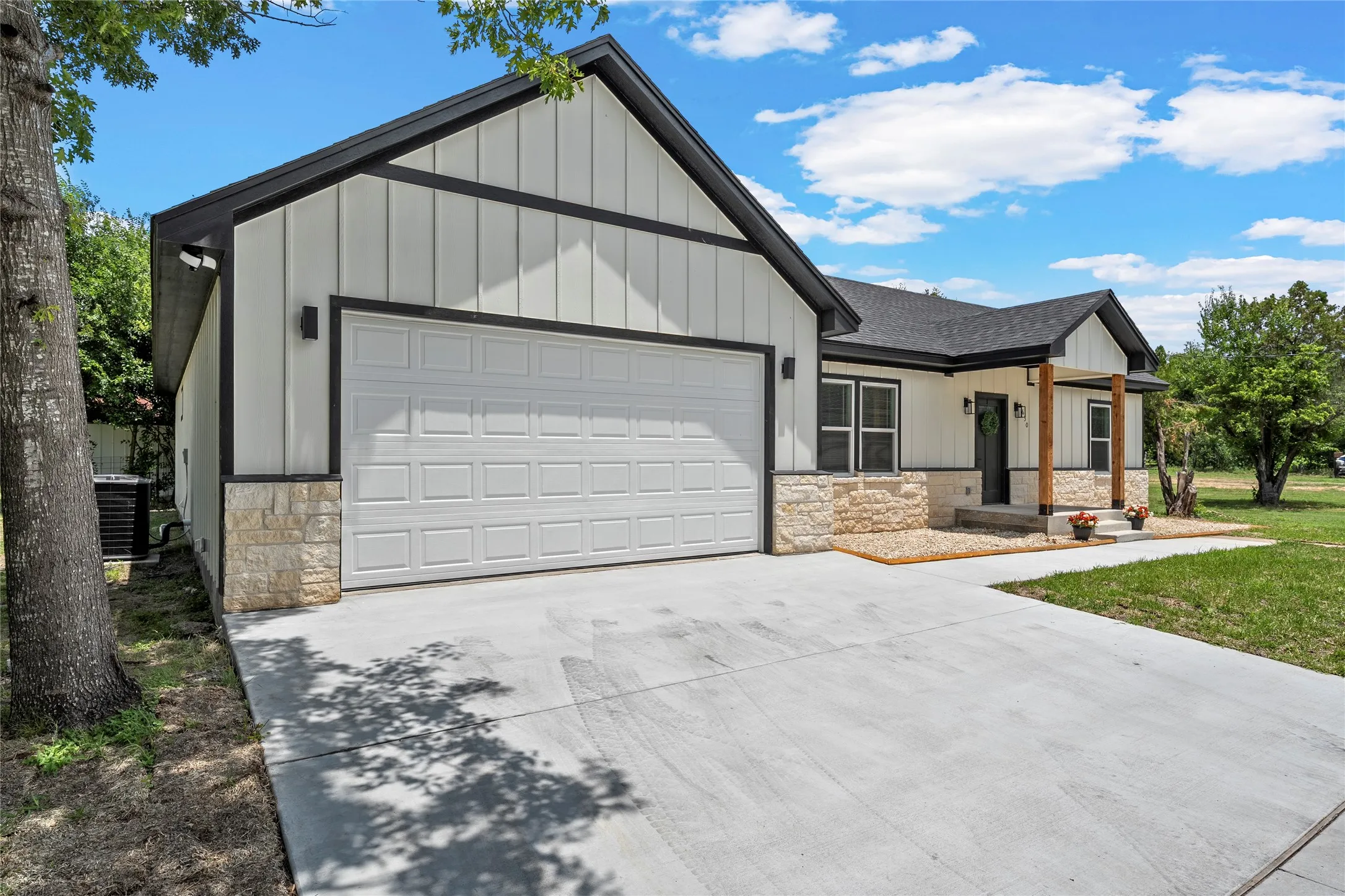 Modern farmhouse exterior with white vertical siding, black trim accents, a two-car garage, stone veneer skirting, and a concrete driveway