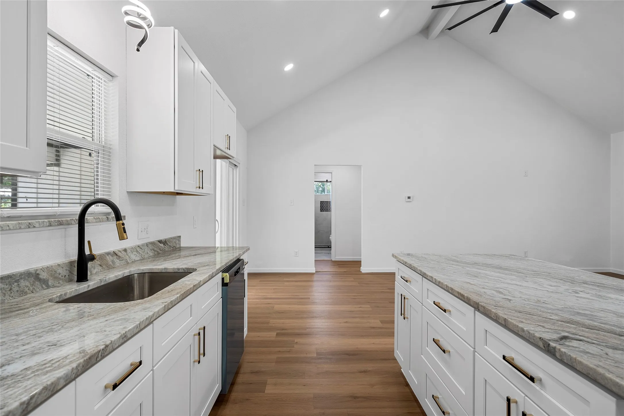Kitchen featuring white shaker cabinetry, light-toned stone countertops, a stainless steel sink with a matte black and gold-tone faucet, and wood-finish flooring