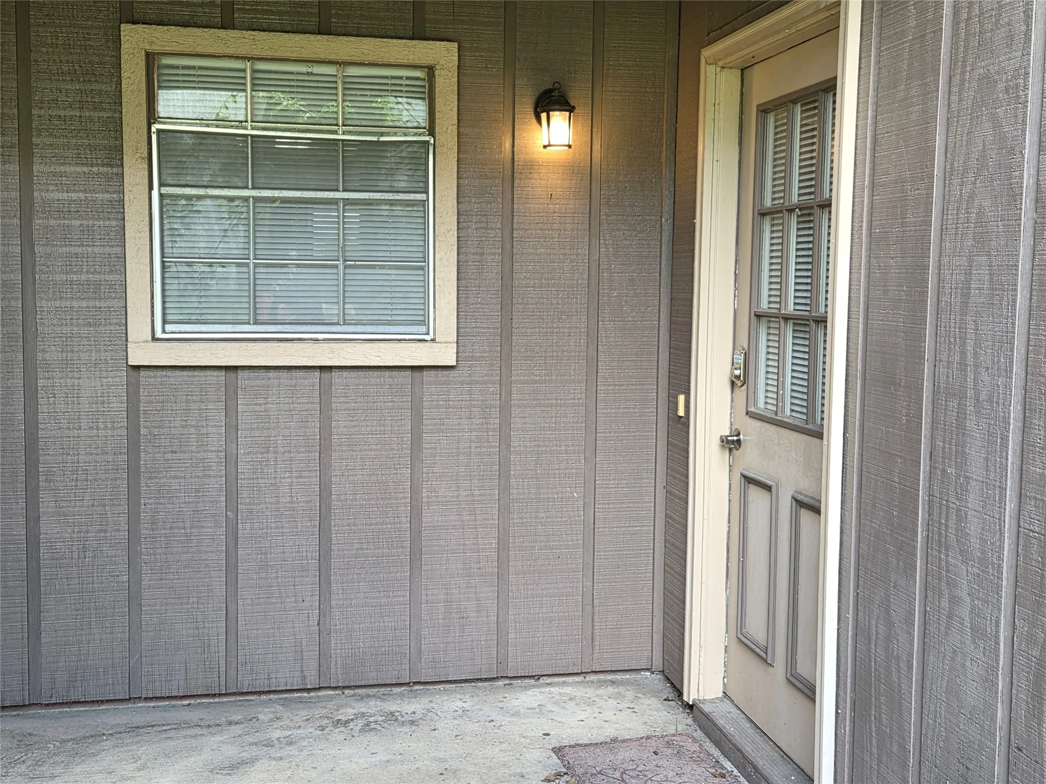 Exterior featuring vertical board-and-batten style siding, a multi-pane window with blinds, a wall-mounted light fixture, and a front entry door with a multi-pane glass insert