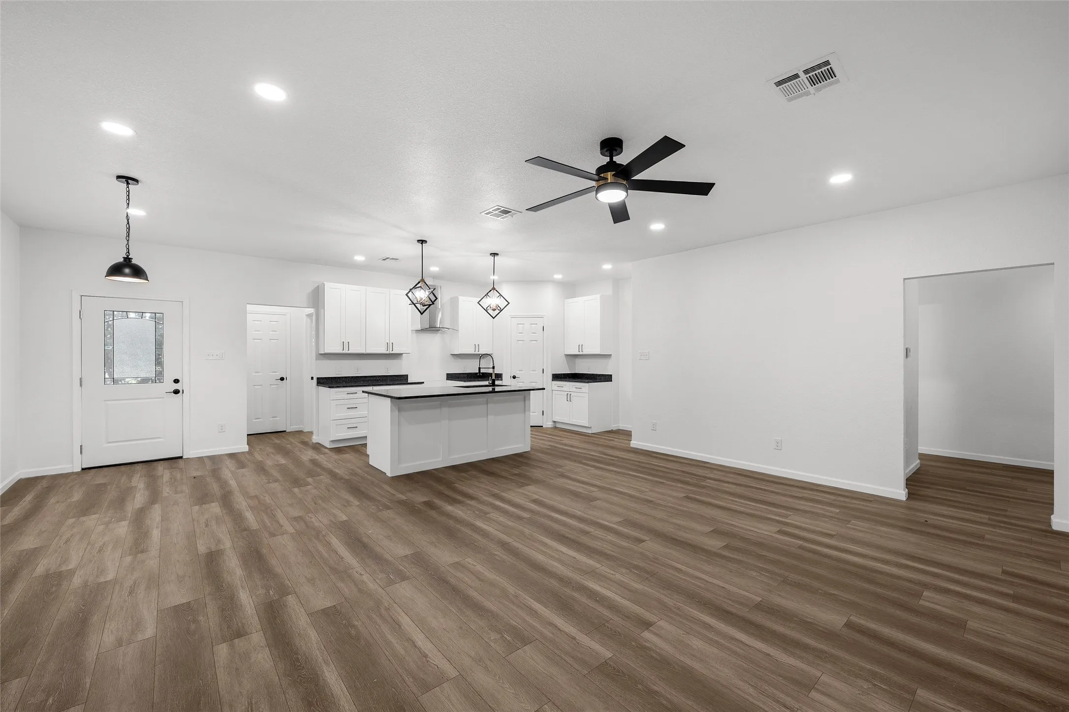 Open-concept living space featuring wood-finish flooring, a modern kitchen island with seating, white shaker cabinetry, dark countertops, and contemporary pendant lighting