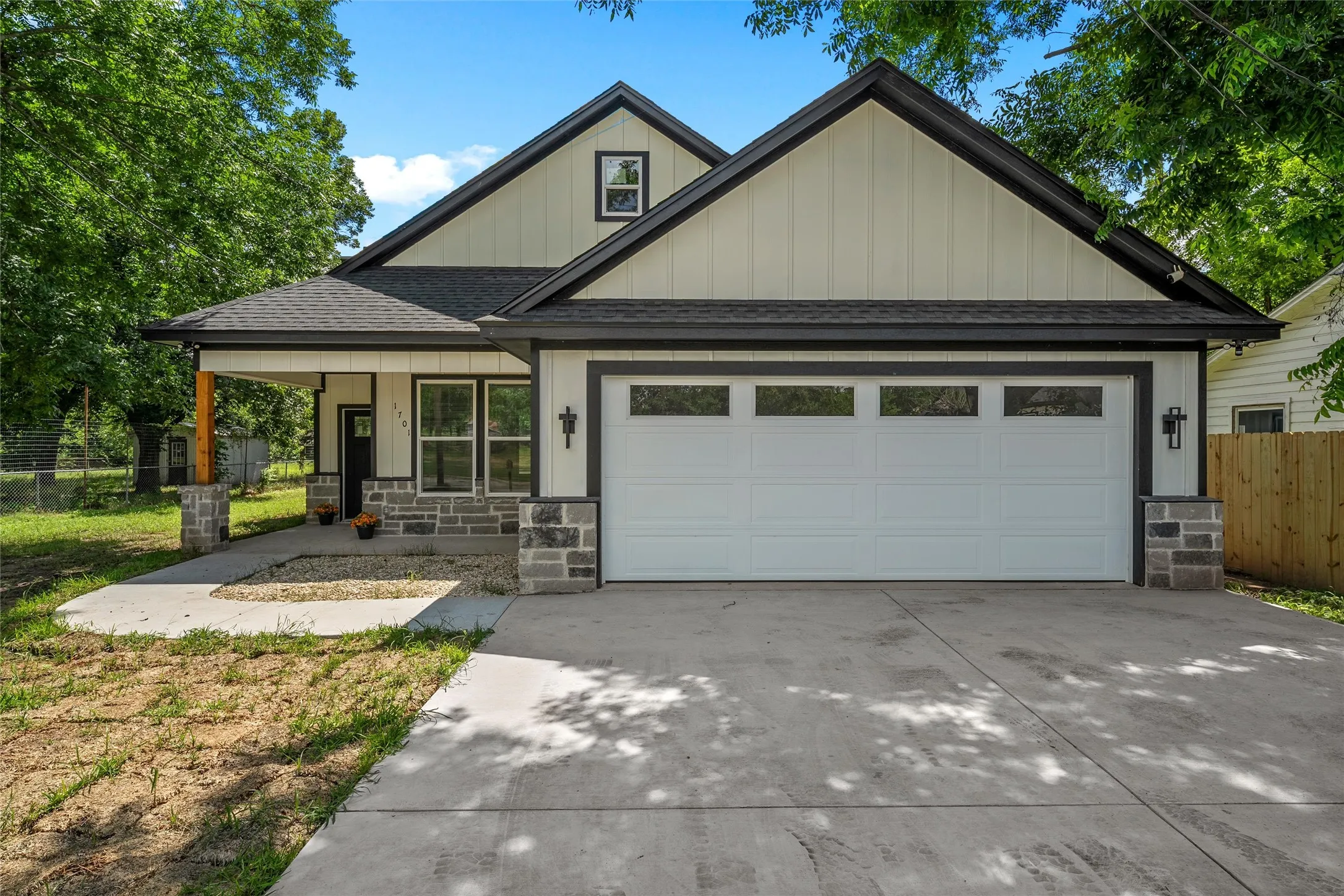 Contemporary exterior featuring light vertical siding, dark trim, a two-car garage with transom windows, stone veneer accents, and a covered front porch with a wood beam post