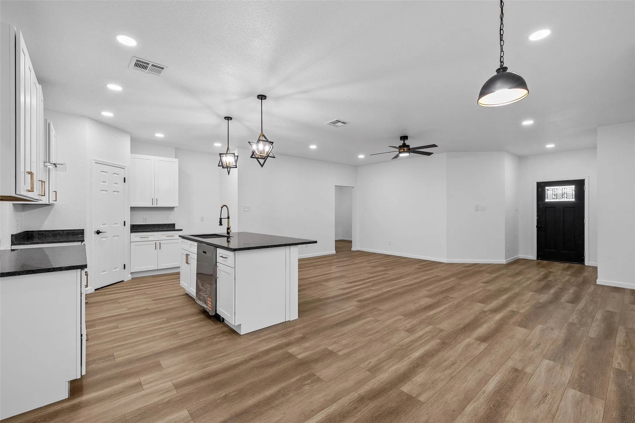 Open-concept living space featuring a contemporary kitchen island with a built-in sink and dishwasher, white cabinetry with gold-tone hardware, dark countertops, and wood-finish flooring throughout