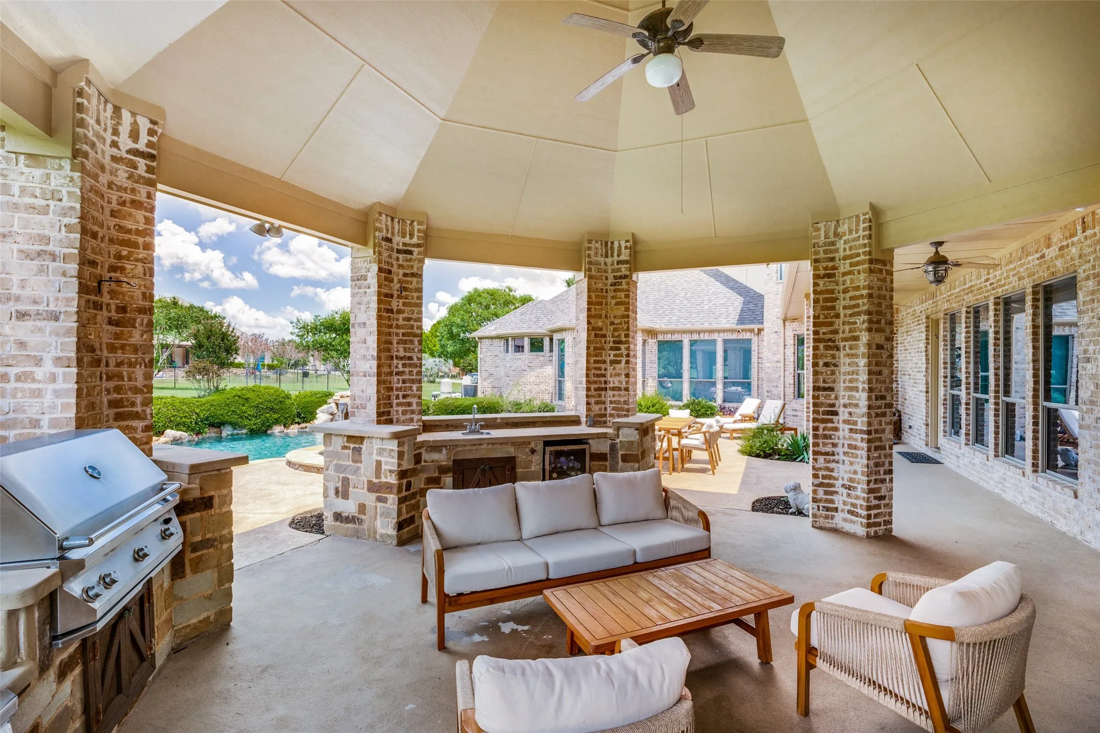 Expansive covered patio featuring brick and stone accent columns, a built-in outdoor kitchen with grill and sink, high ceilings with ceiling fans, and concrete flooring
