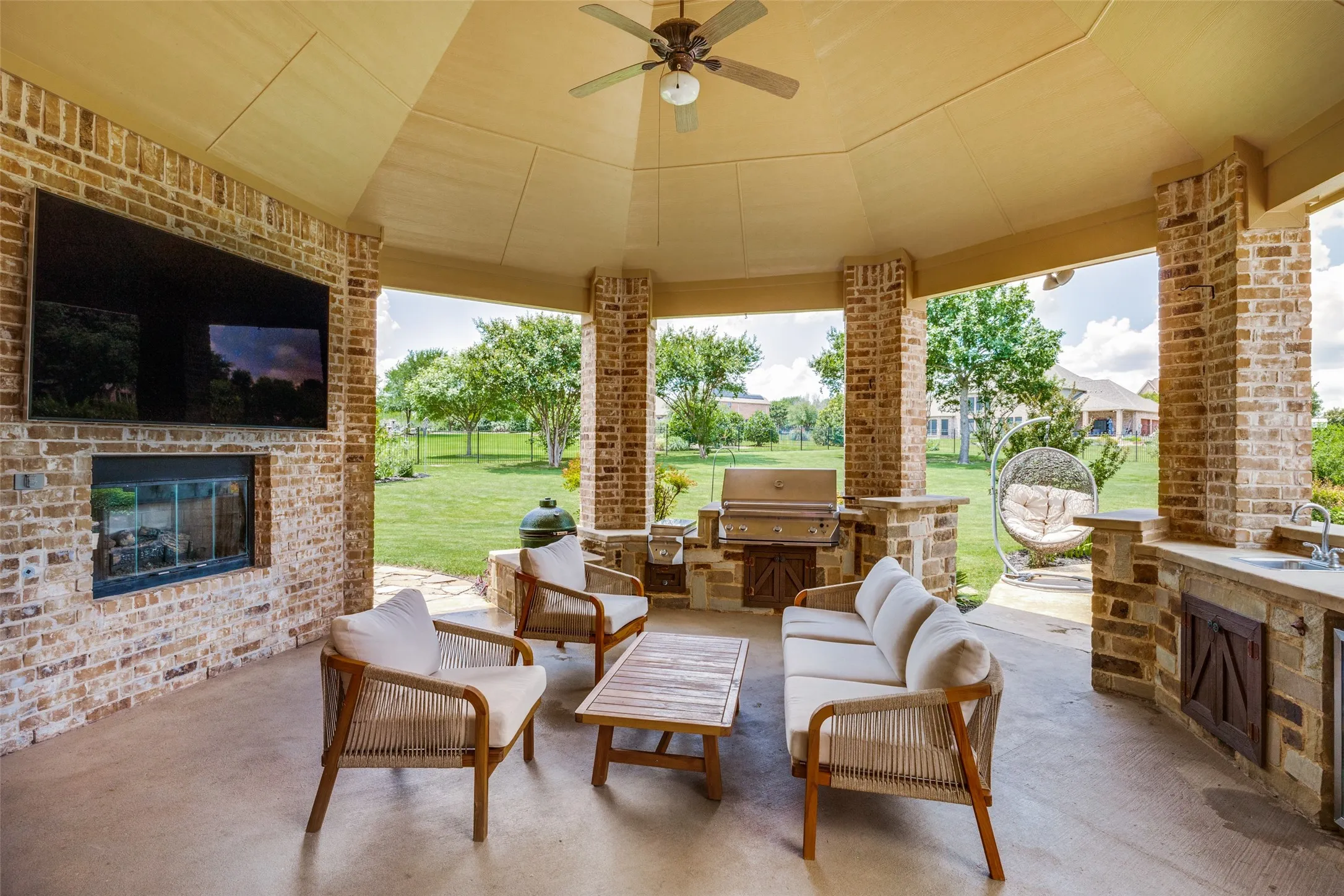 Expansive covered patio featuring brick columns, a built-in outdoor kitchen with grill and sink, a ceiling fan, and a mounted television above an outdoor fireplace