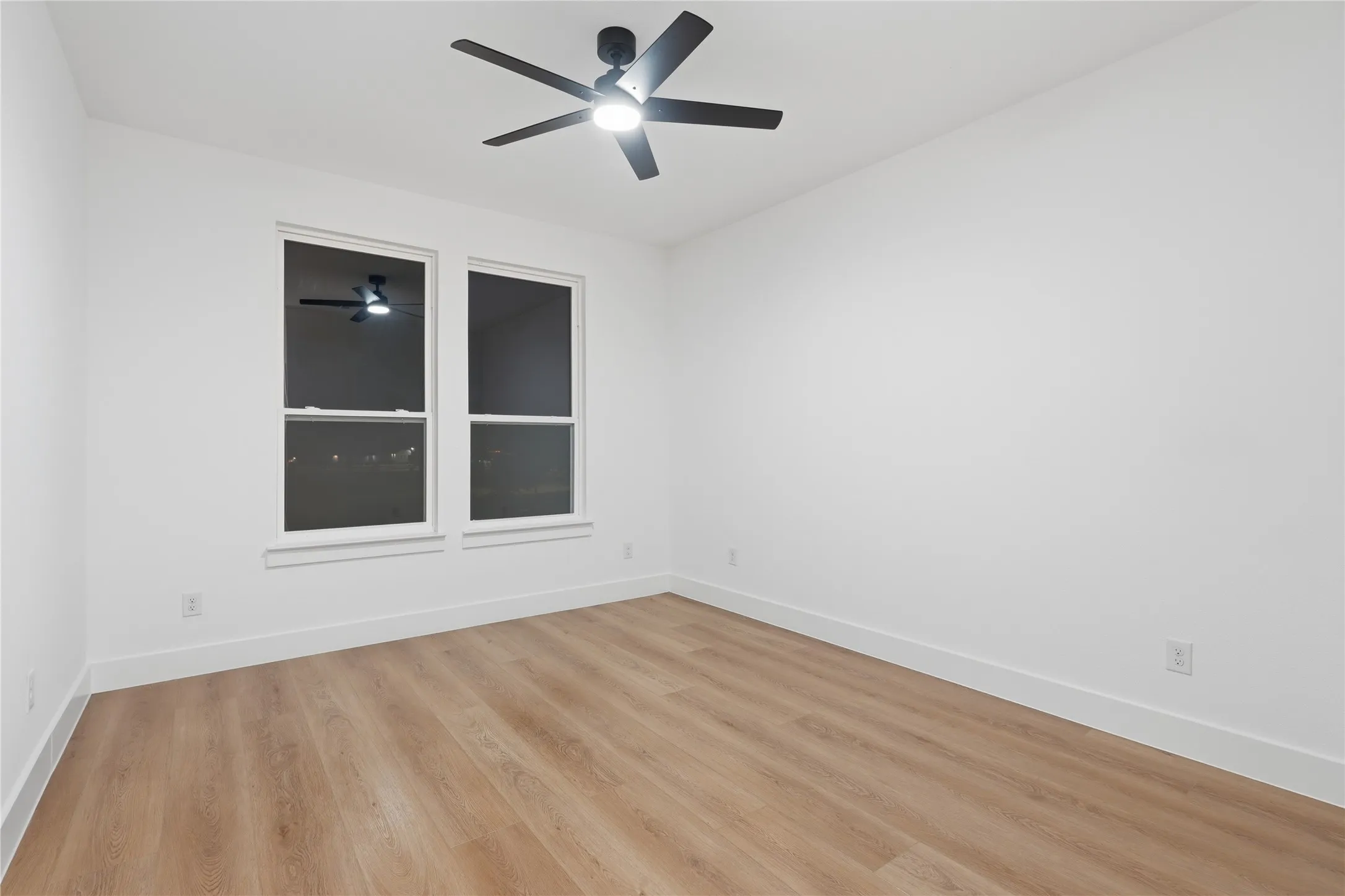 Bright room featuring two large windows, wood-finish flooring, white baseboards, and a modern ceiling fan with integrated lighting
