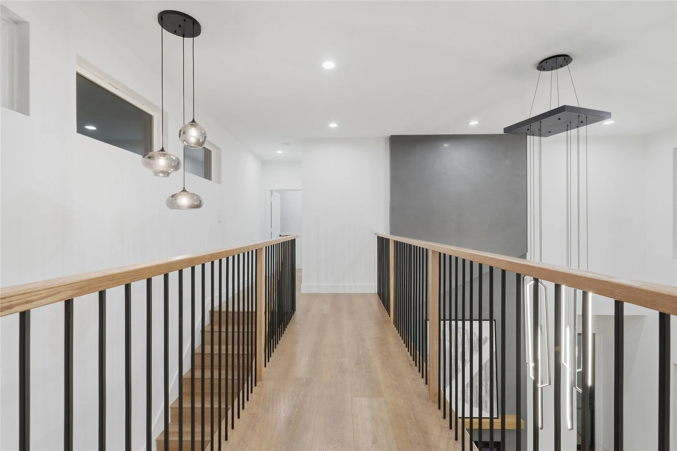 Upper-level hallway featuring wood-finish flooring, contemporary lighting fixtures, a light wood handrail with black iron balusters, recessed ceiling lights, and transom windows