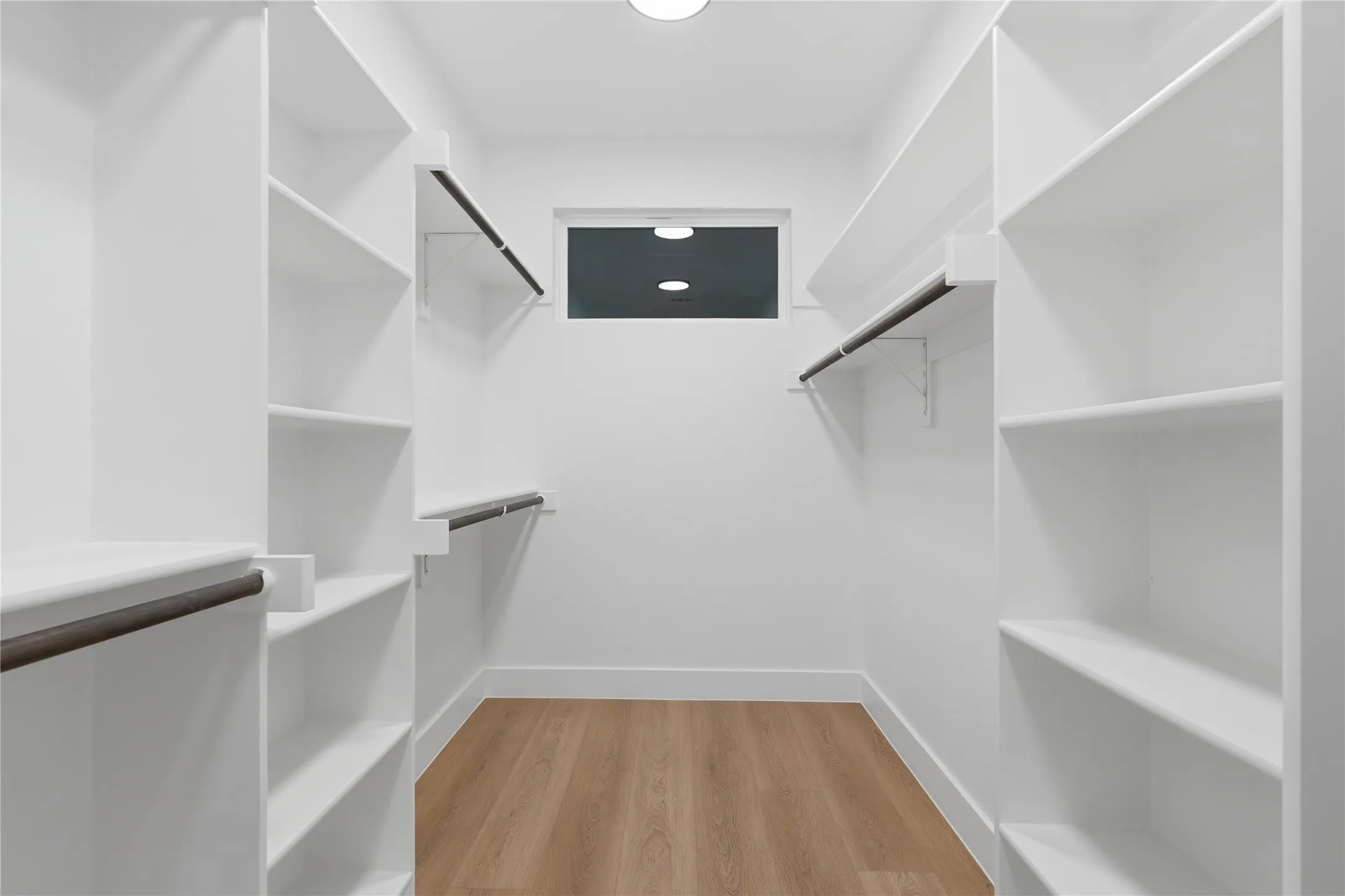 Walk-in closet featuring extensive white shelving, multiple hanging rods, a transom window, wood-finish flooring, and bright overhead lighting