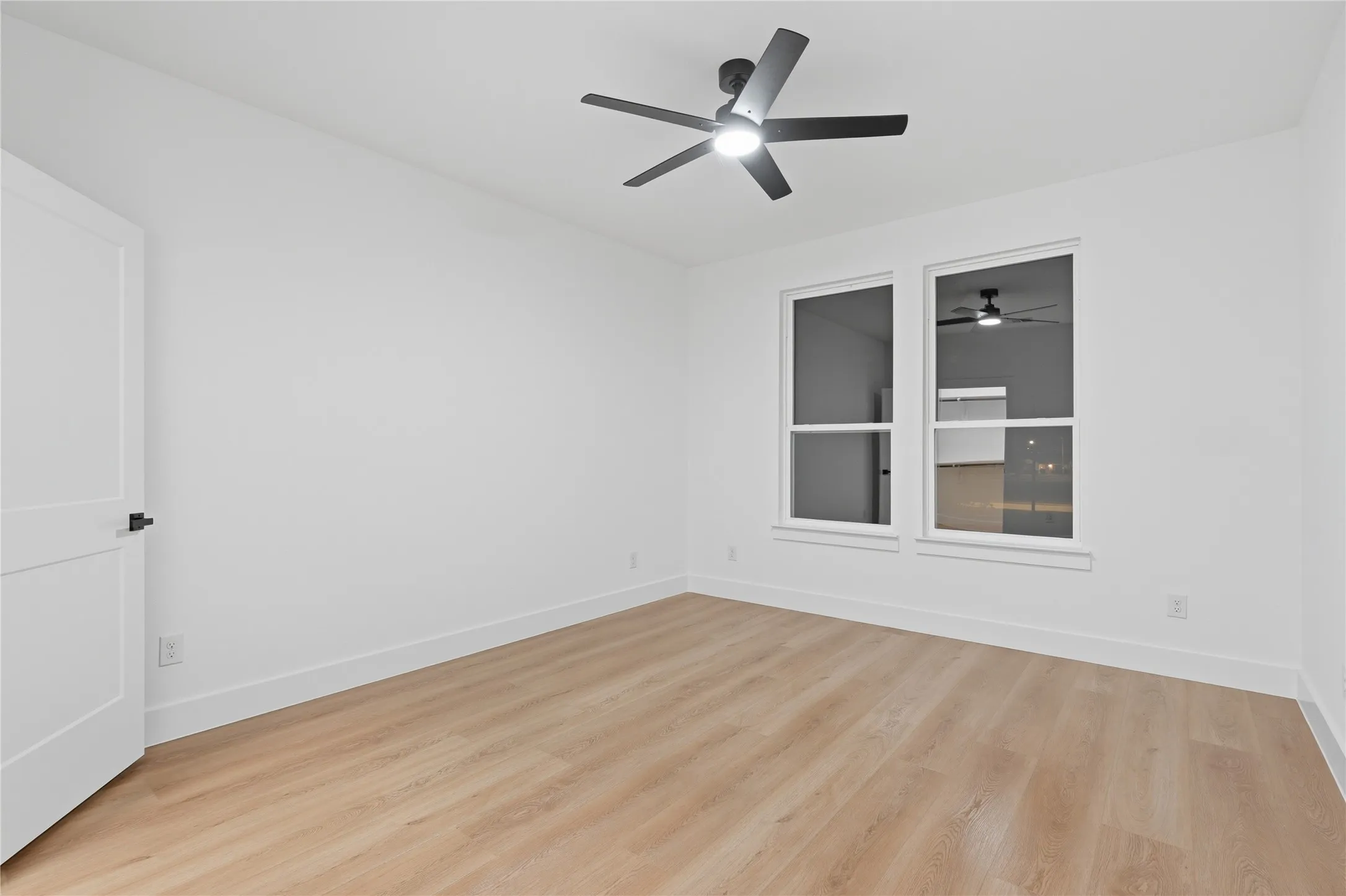 Bright room featuring two large windows, a ceiling fan with integrated lighting, wood-finish flooring, and a contemporary white door with matte black hardware
