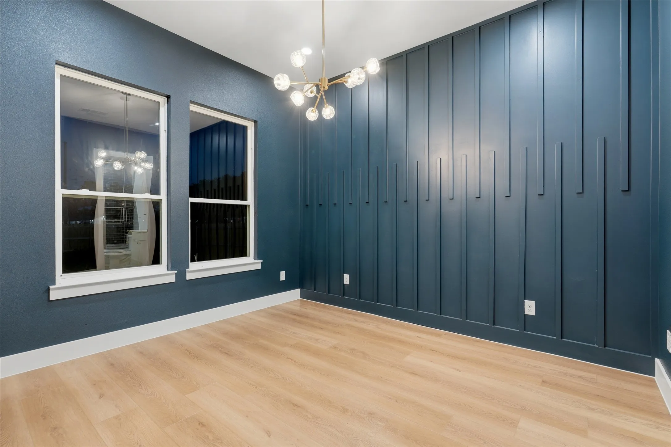 Room featuring a decorative accent wall with vertical paneling, two double-hung windows, wood-finish flooring, and a modern chandelier with spherical lights
