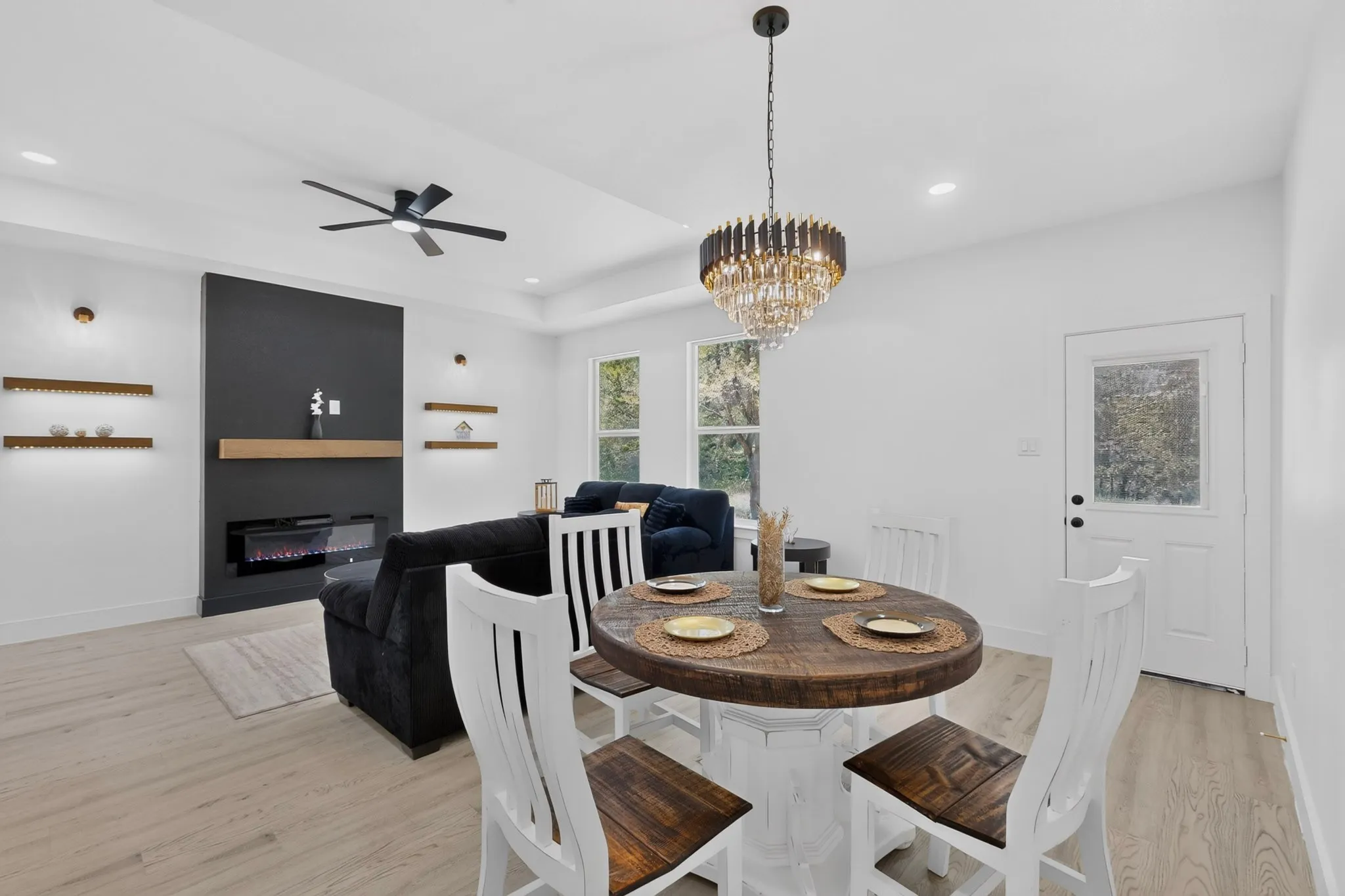 Open-concept living space featuring wood-finish flooring, a modern electric fireplace with a dark accent wall and light wood mantel, and illuminated floating shelves